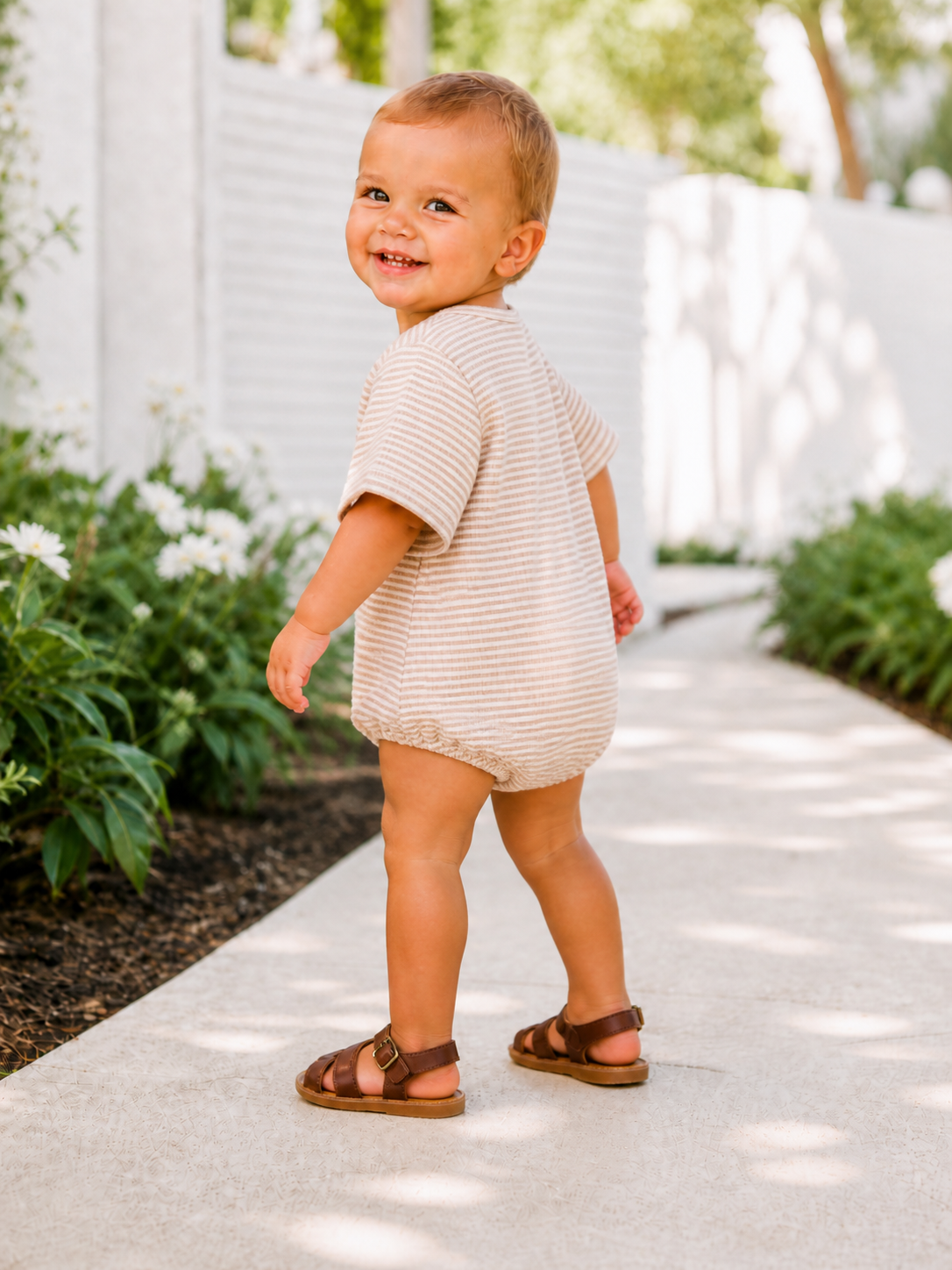 Smiling toddler in a striped outfit walking on a path surrounded by flowers and greenery.