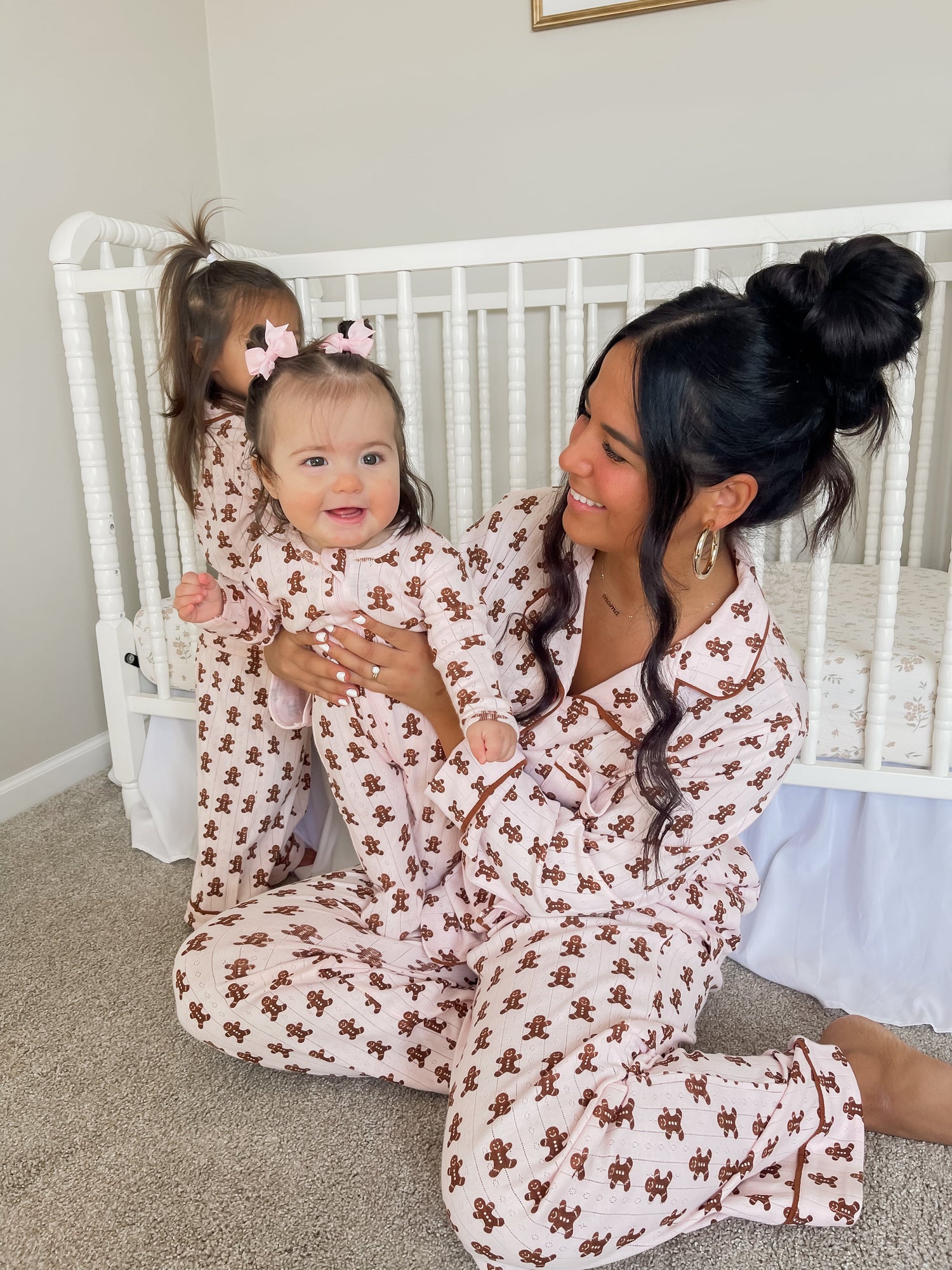 Mother and baby wearing matching pink gingerbread-themed pajamas, seated near a crib, with a playful atmosphere.
