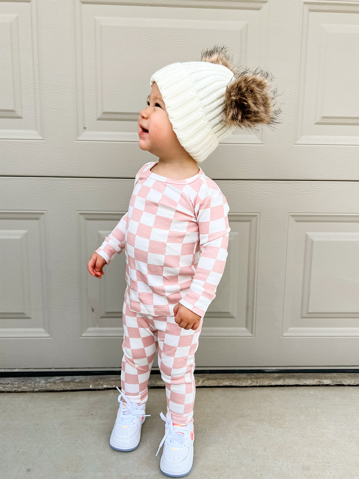 Toddler in a pink and white checkered outfit with a fluffy hat, standing outside near a garage door.