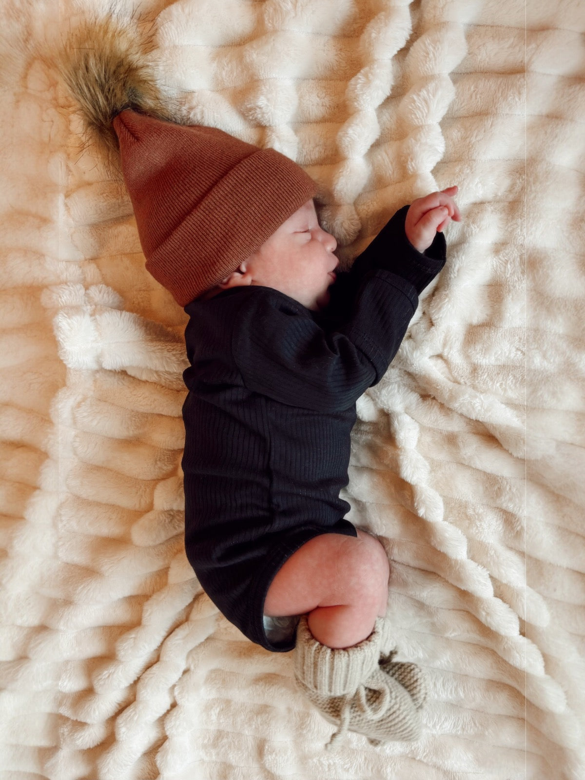 Newborn peacefully sleeping on a plush blanket, wearing a brown beanie and cozy outfit with knit booties.