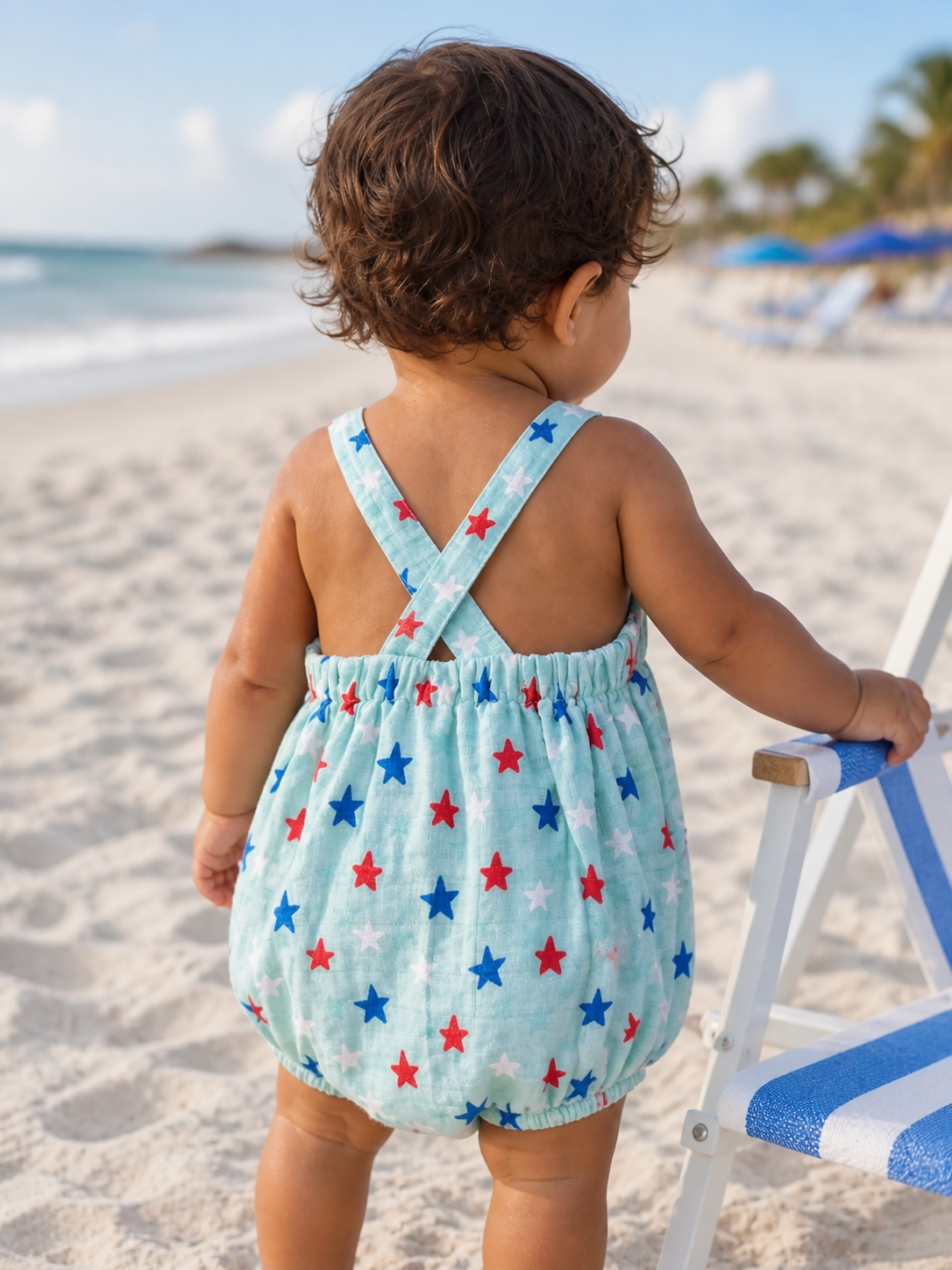 Child in a star-patterned romper stands on the beach, facing the ocean, with beach chairs in the background.