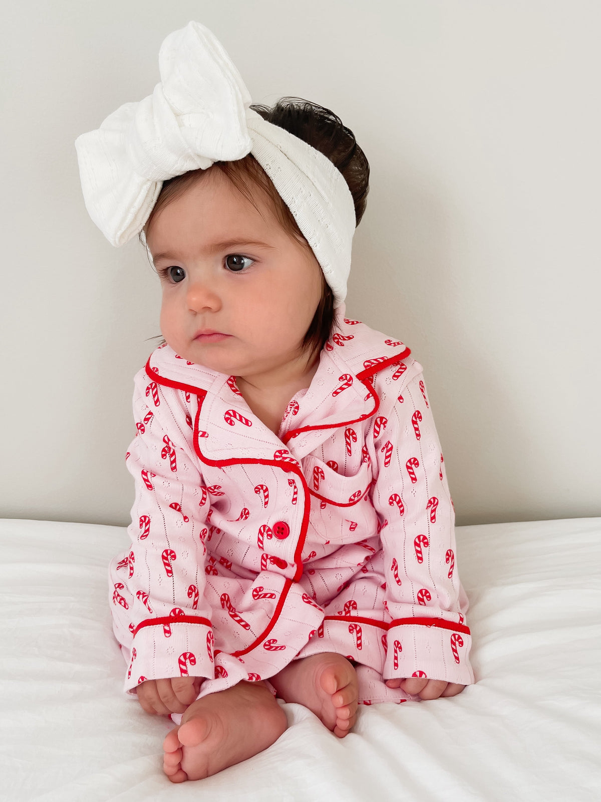 Baby in candy cane pajamas and a headband sitting on a bed, looking thoughtful against a light background.