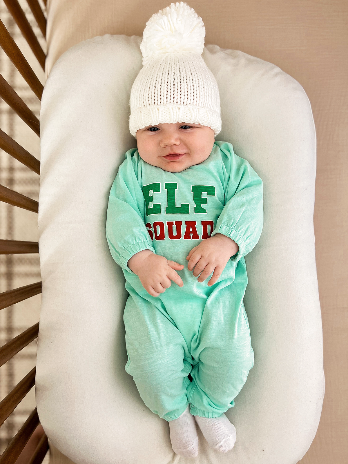Smiling baby in an aqua outfit with “ELF SQUAD” text and white knitted hat, resting on a soft pillow.