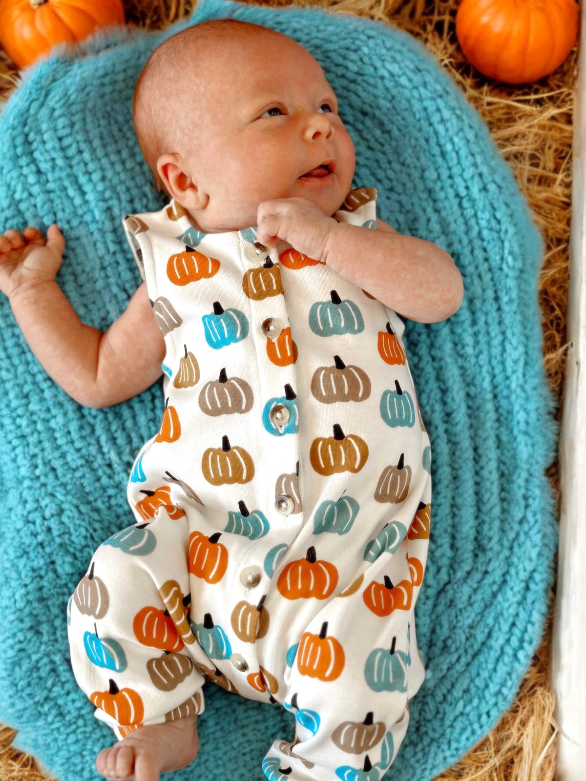 Baby in a pumpkin-patterned romper lies on a blue blanket surrounded by pumpkins and hay.