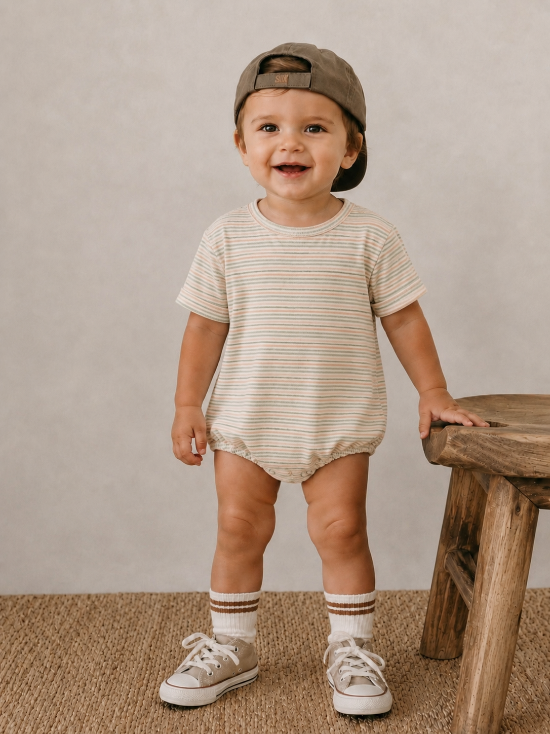 Smiling toddler in striped outfit and cap, standing beside a wooden stool on a textured surface.