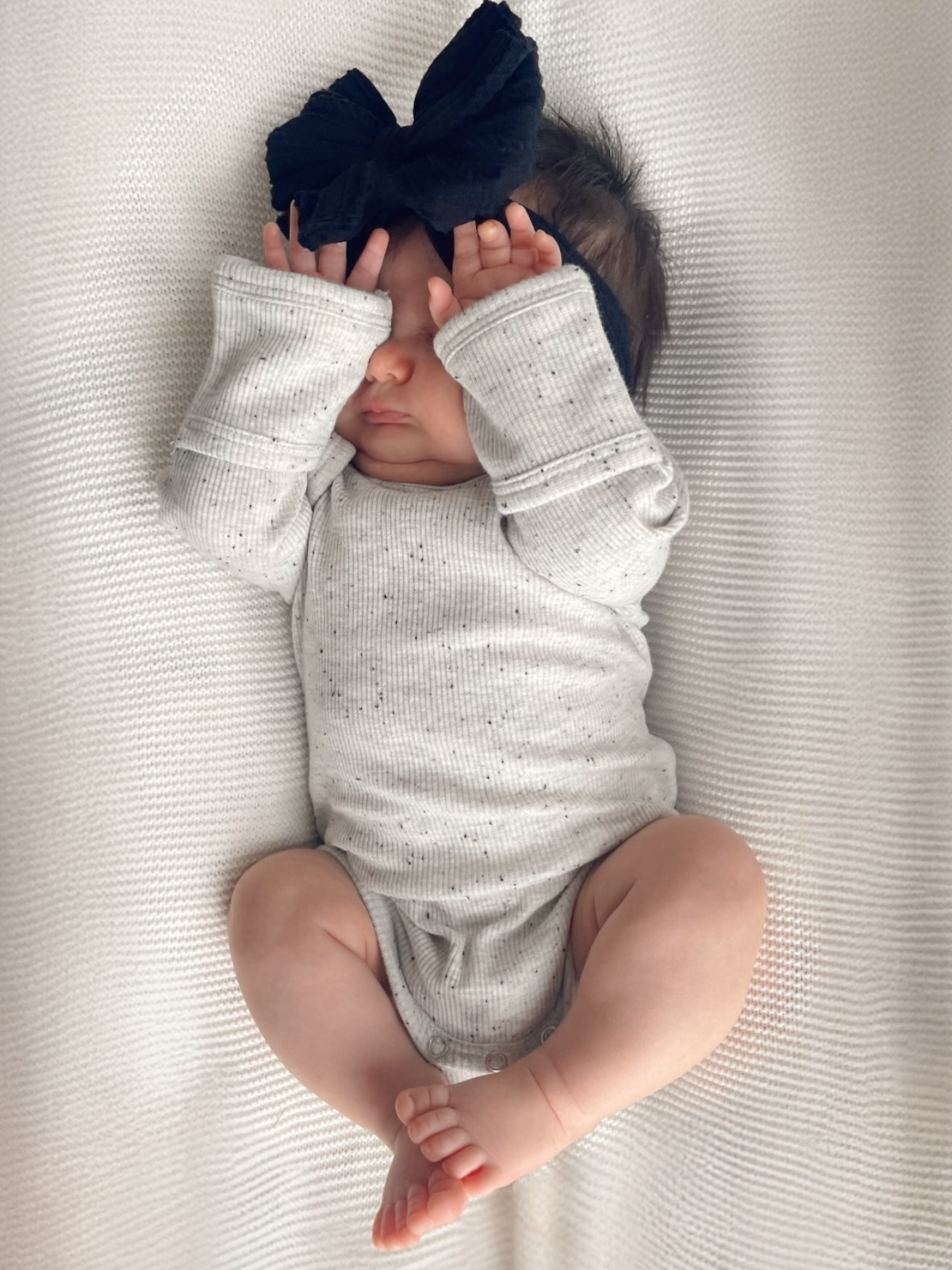 Infant wearing a gray onesie and a large black bow, playfully covering their eyes with hands on a soft white background.