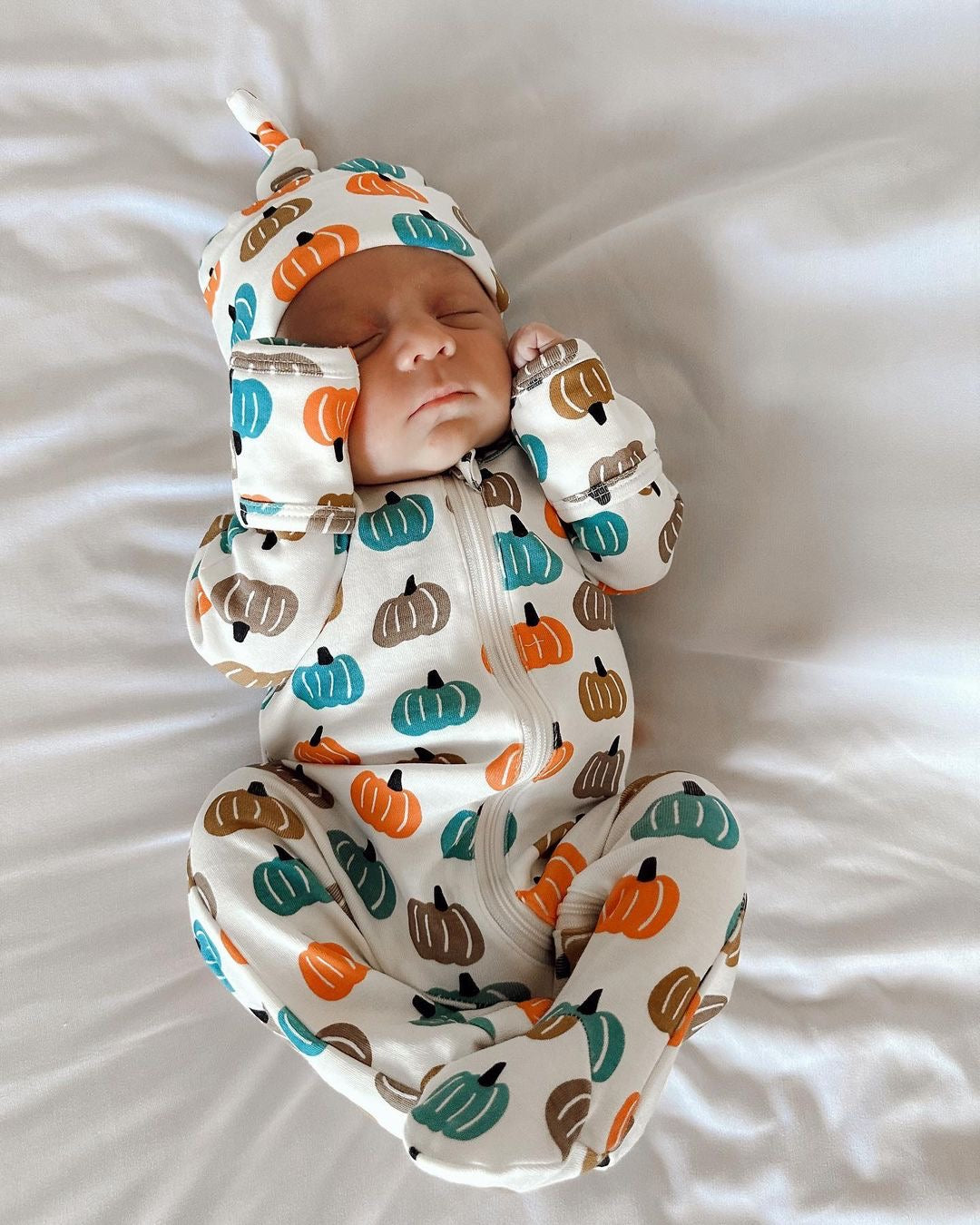 Newborn baby in a pumpkin-patterned onesie and hat, sleeping on a white blanket.