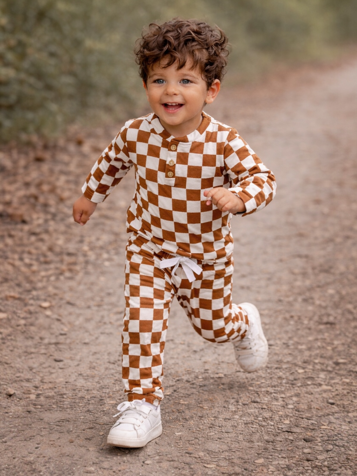 Smiling child in brown and white checkered outfit running on a dirt path surrounded by greenery.
