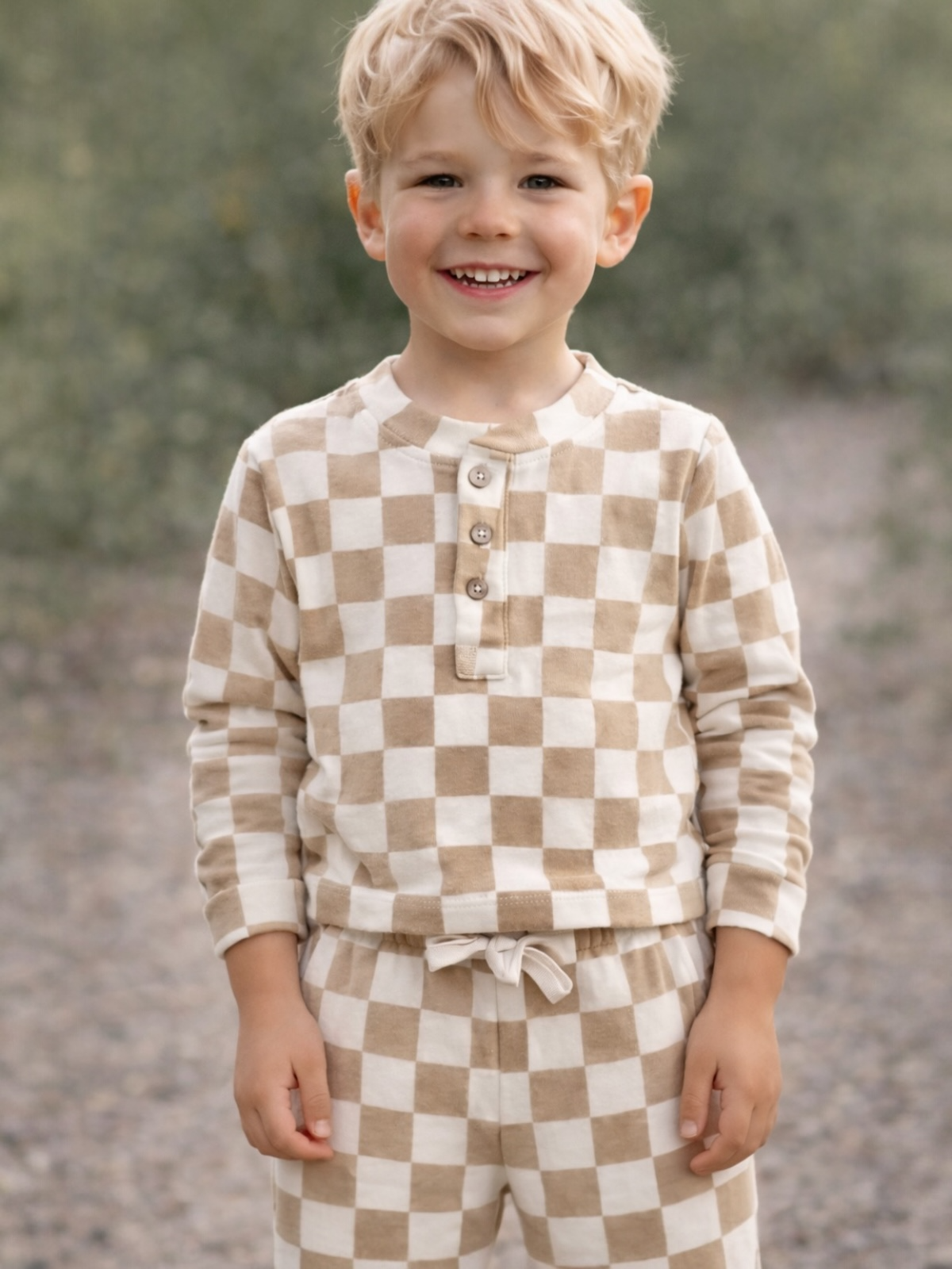 Smiling boy in checkered pajamas standing outdoors, surrounded by greenery.