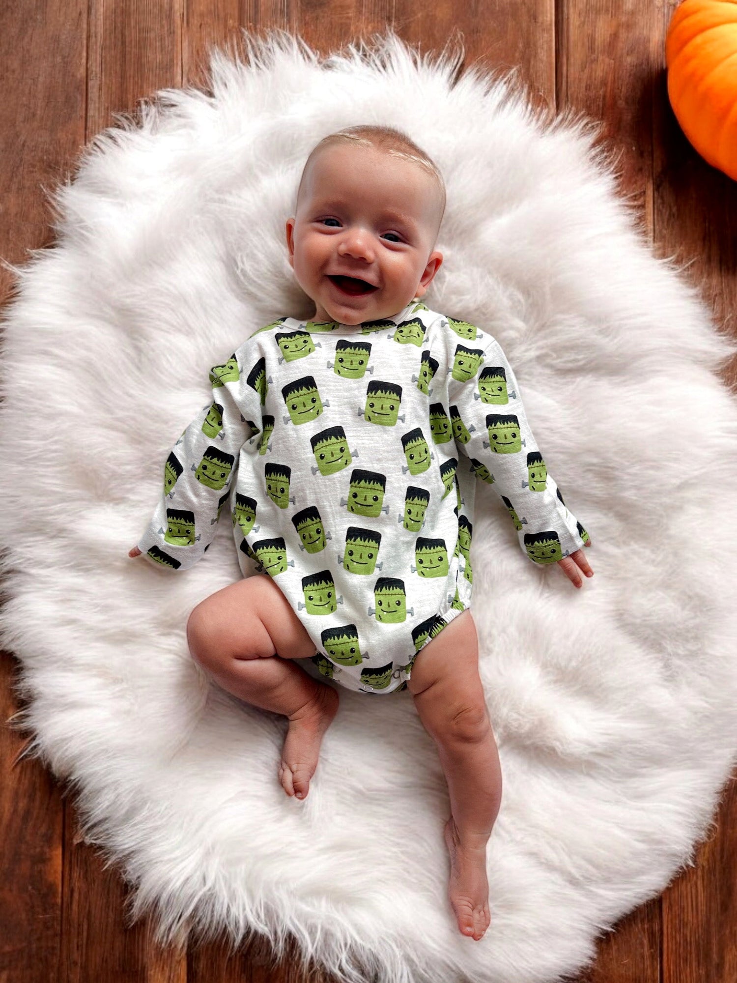 Smiling baby on fluffy white rug, wearing a Frankenstein-themed onesie, surrounded by a cozy autumn setting.