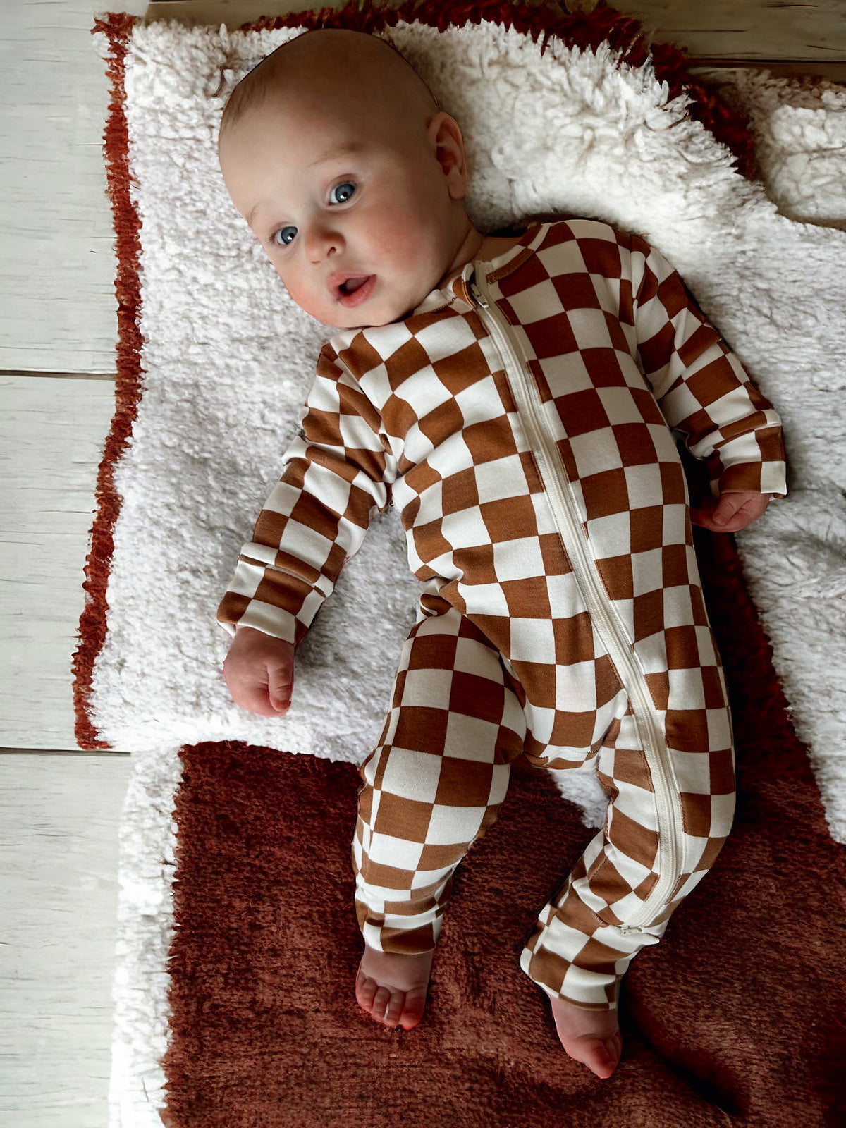 Infant lying on a soft rug, wearing a brown and white checkered zip-up onesie, with a curious expression.