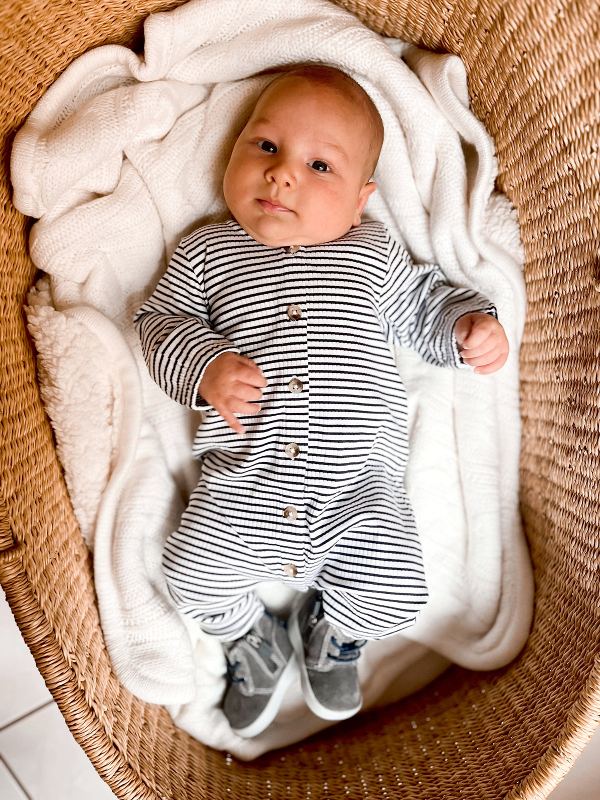 Baby in a striped onesie lying in a woven basket with a cozy blanket, looking at the camera.