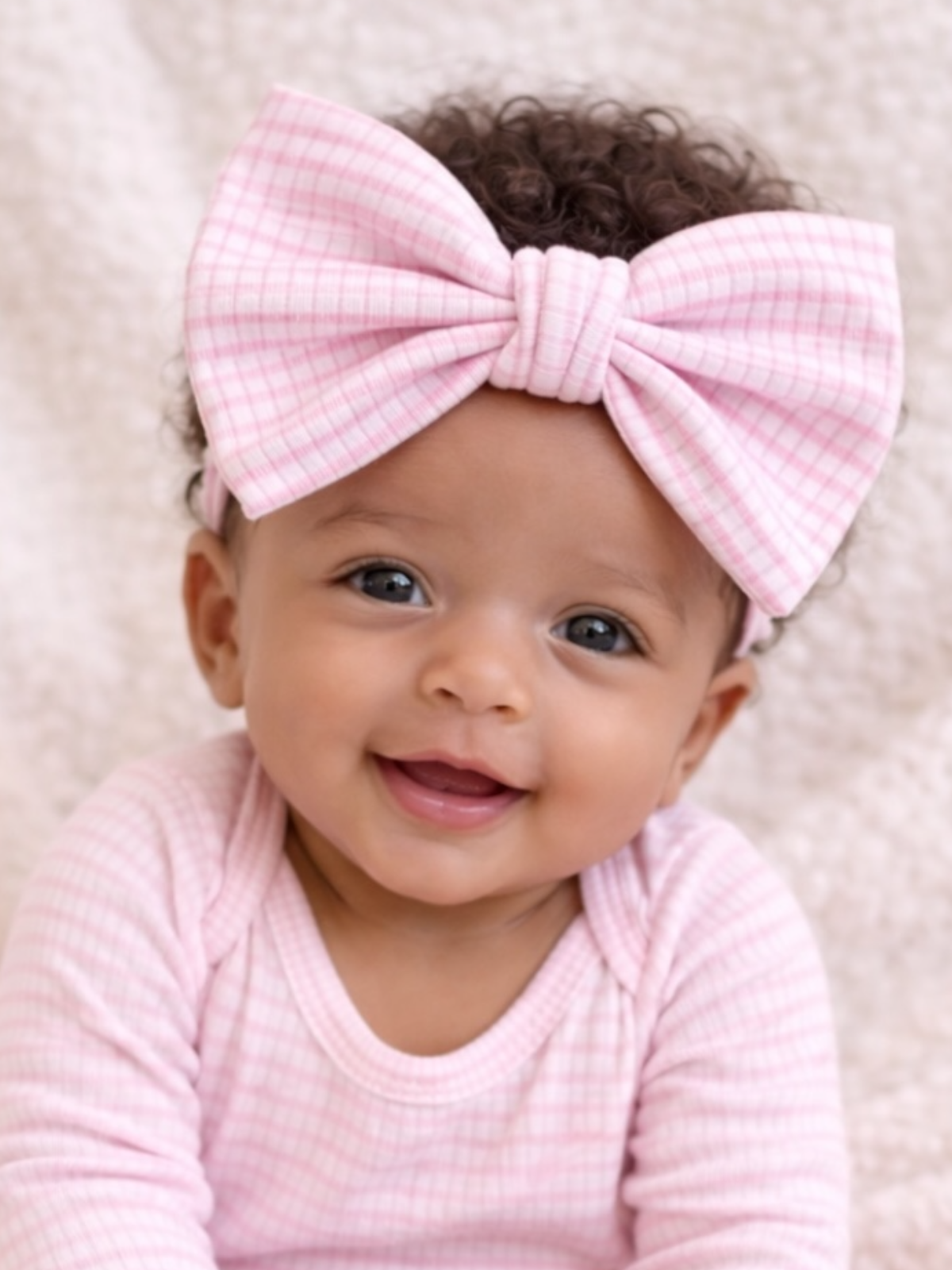 Smiling baby girl with curly hair wearing a pink checkered outfit and a large pink bow headband.
