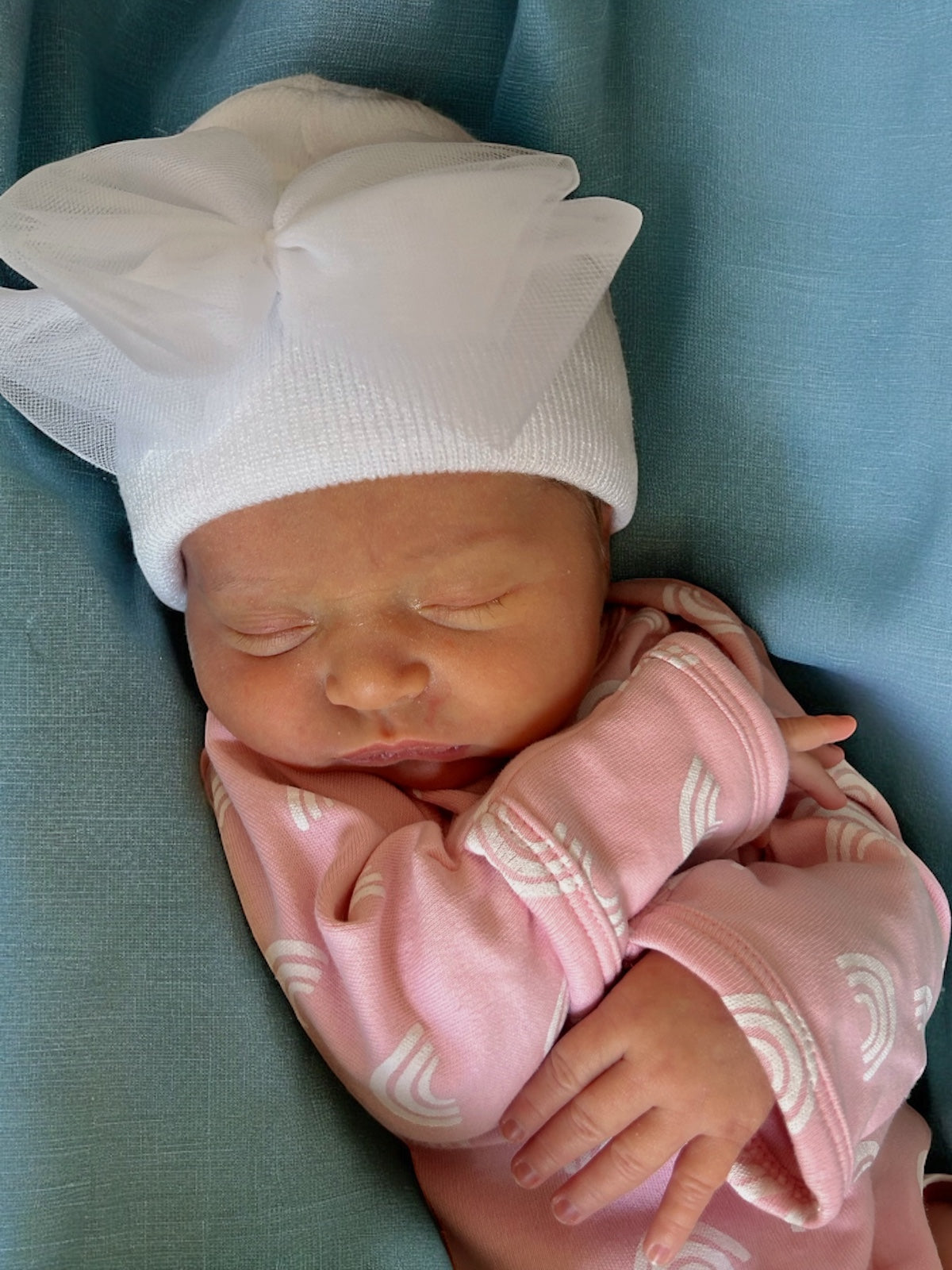 Newborn baby girl with a large white bow, wearing a pink outfit with rainbow patterns, sleeping peacefully on a blue background.