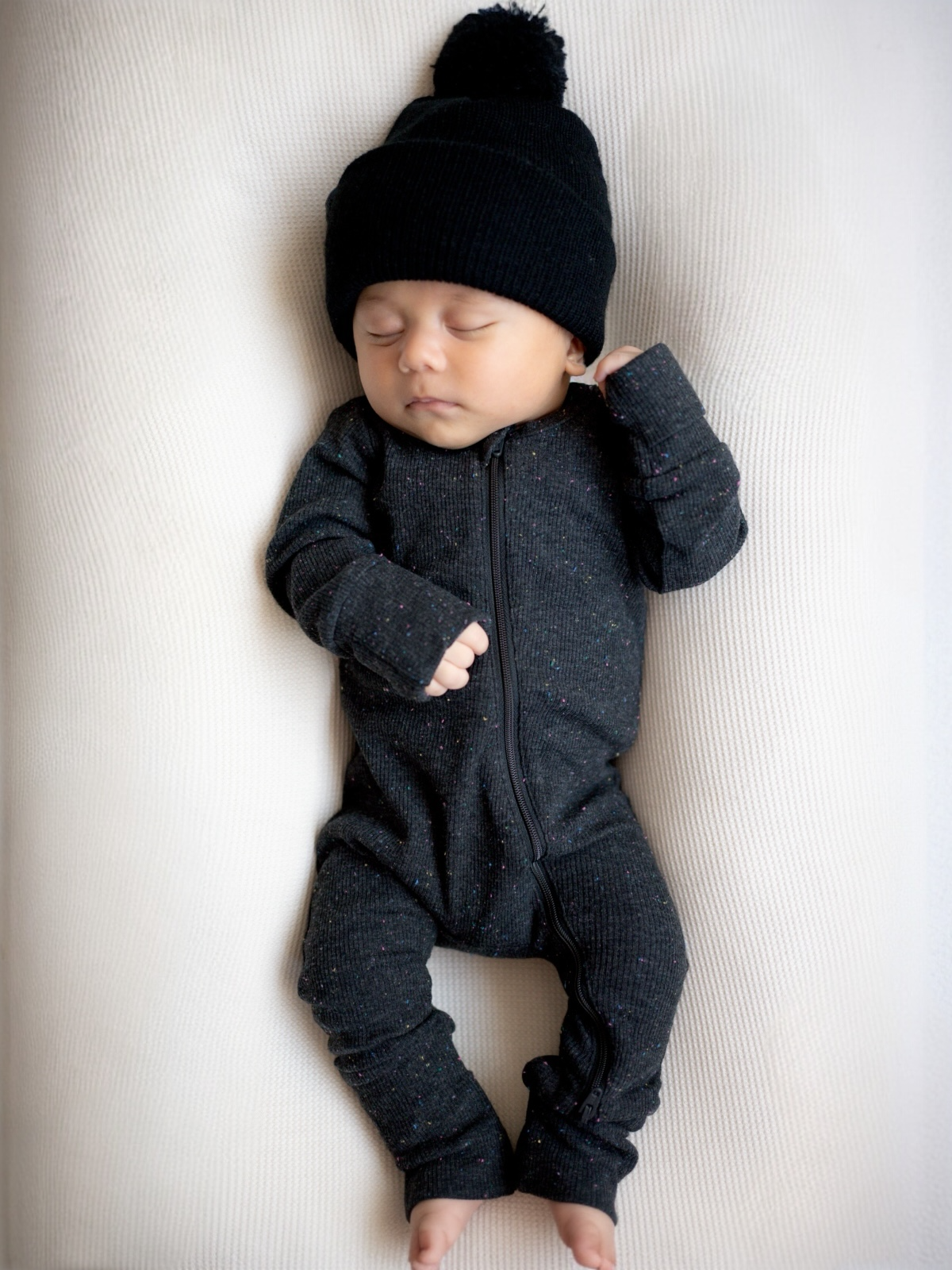 Baby in a black zip-up onesie and pom-pom hat sleeping peacefully on a light-colored surface.