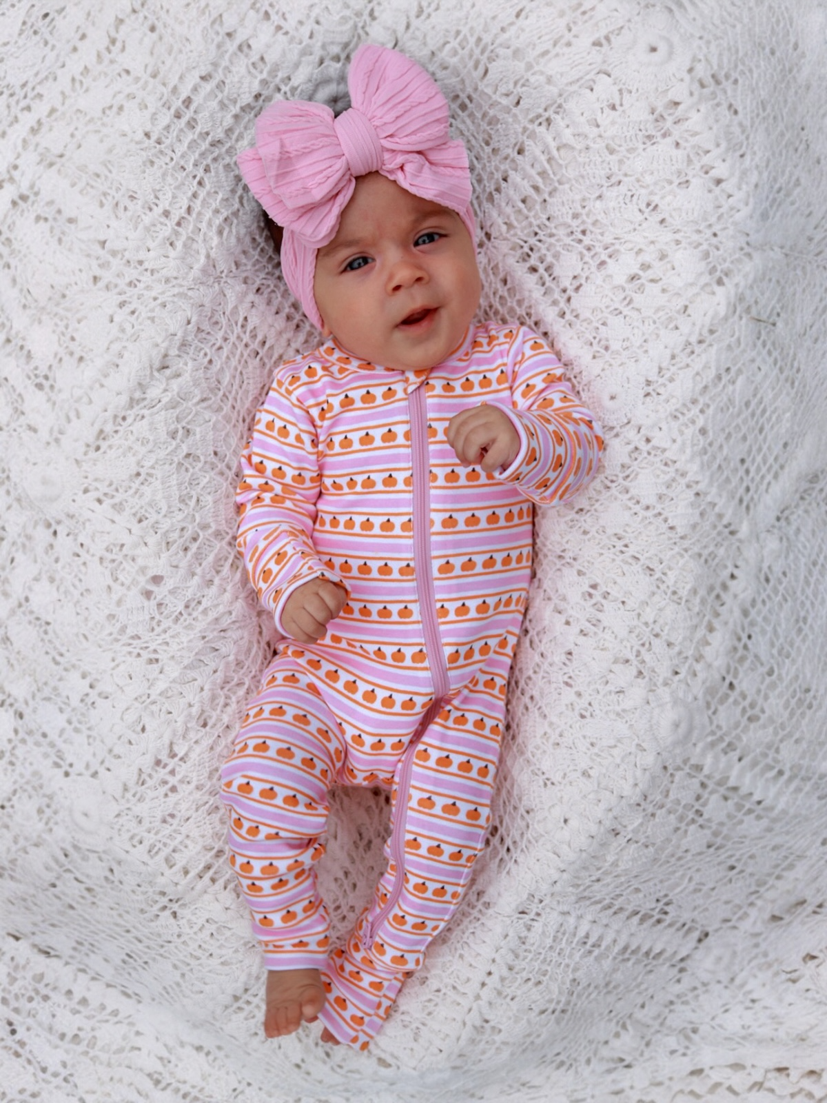 Baby wearing a pink headband and orange-patterned onesie, lying on a white knitted blanket, looking curiously at the camera.