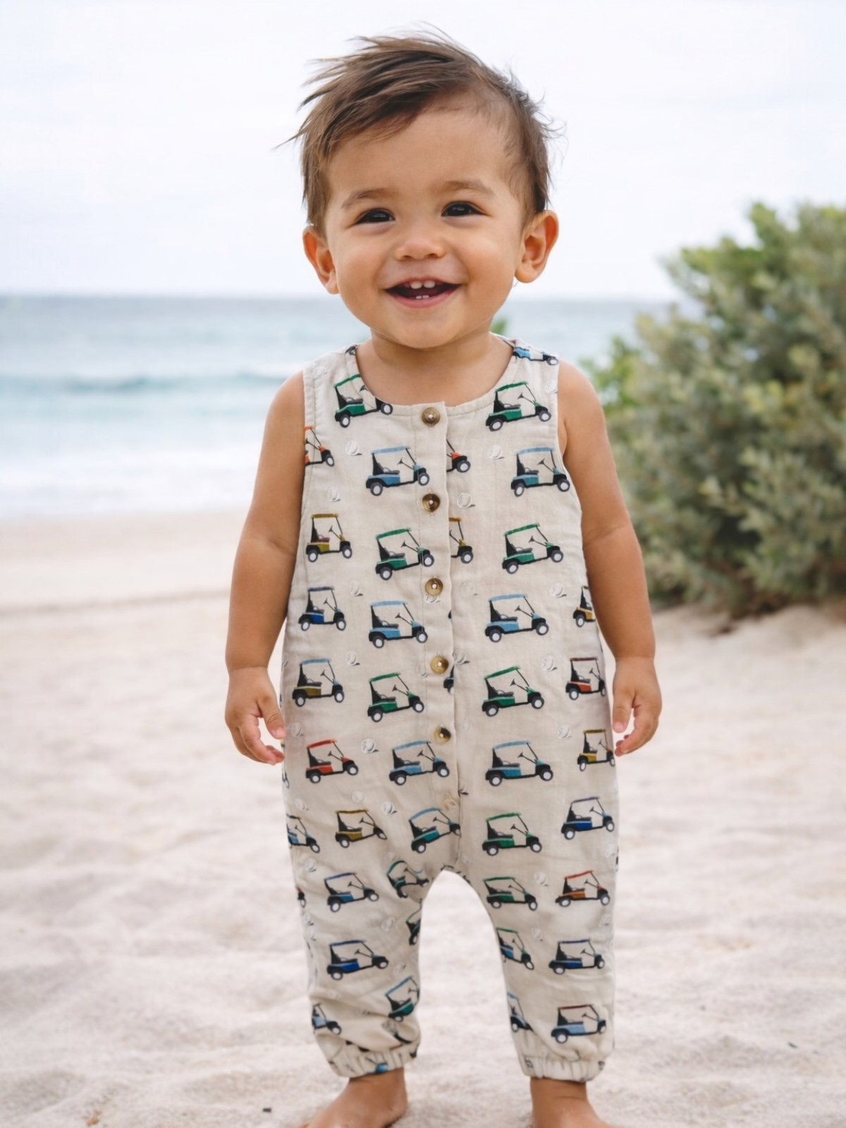 Smiling toddler in a patterned romper stands on the beach, with ocean waves and greenery in the background.