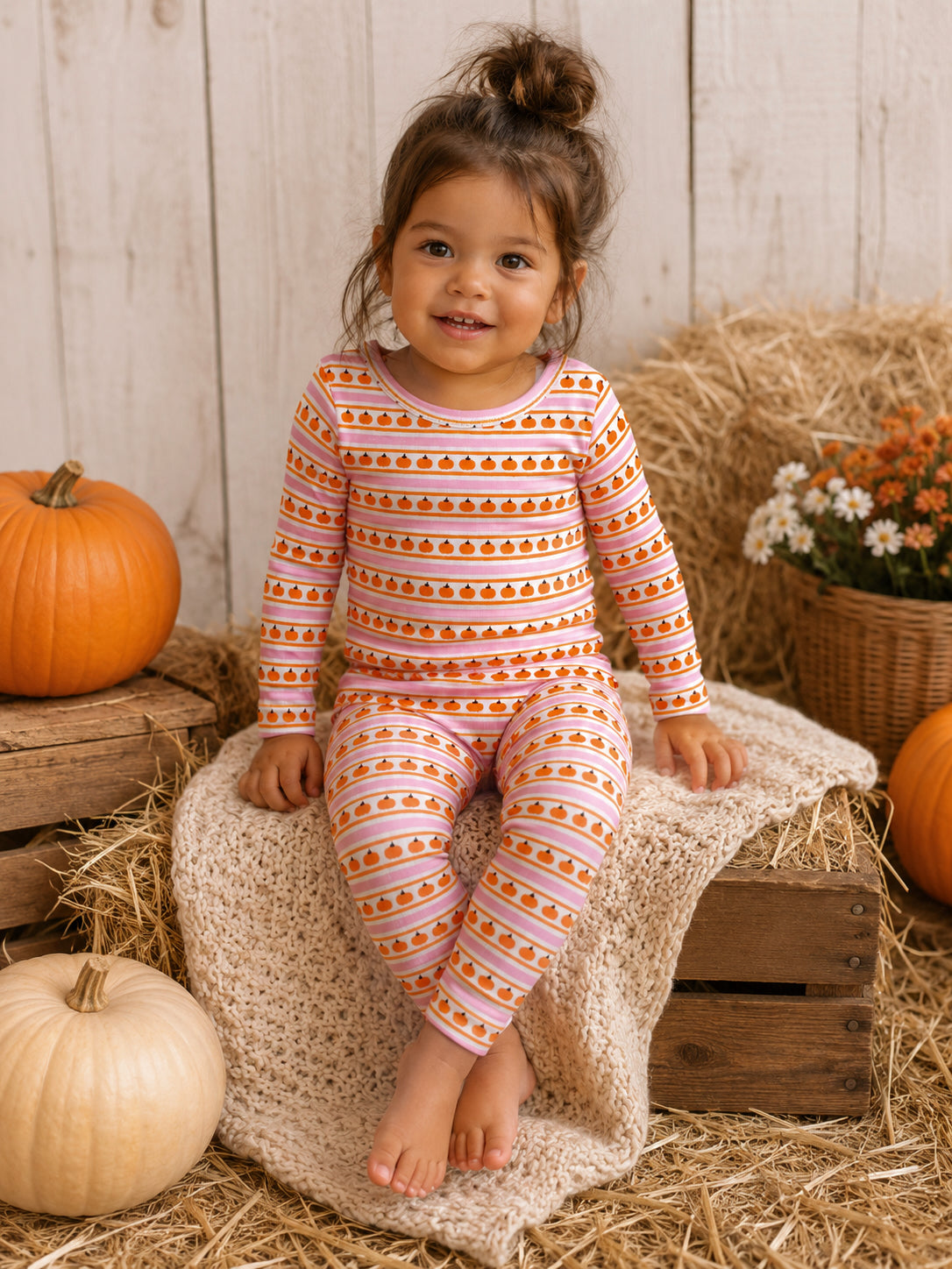 Smiling child in pumpkin-themed pajamas, sitting on straw and a cozy blanket with pumpkins and flowers around.