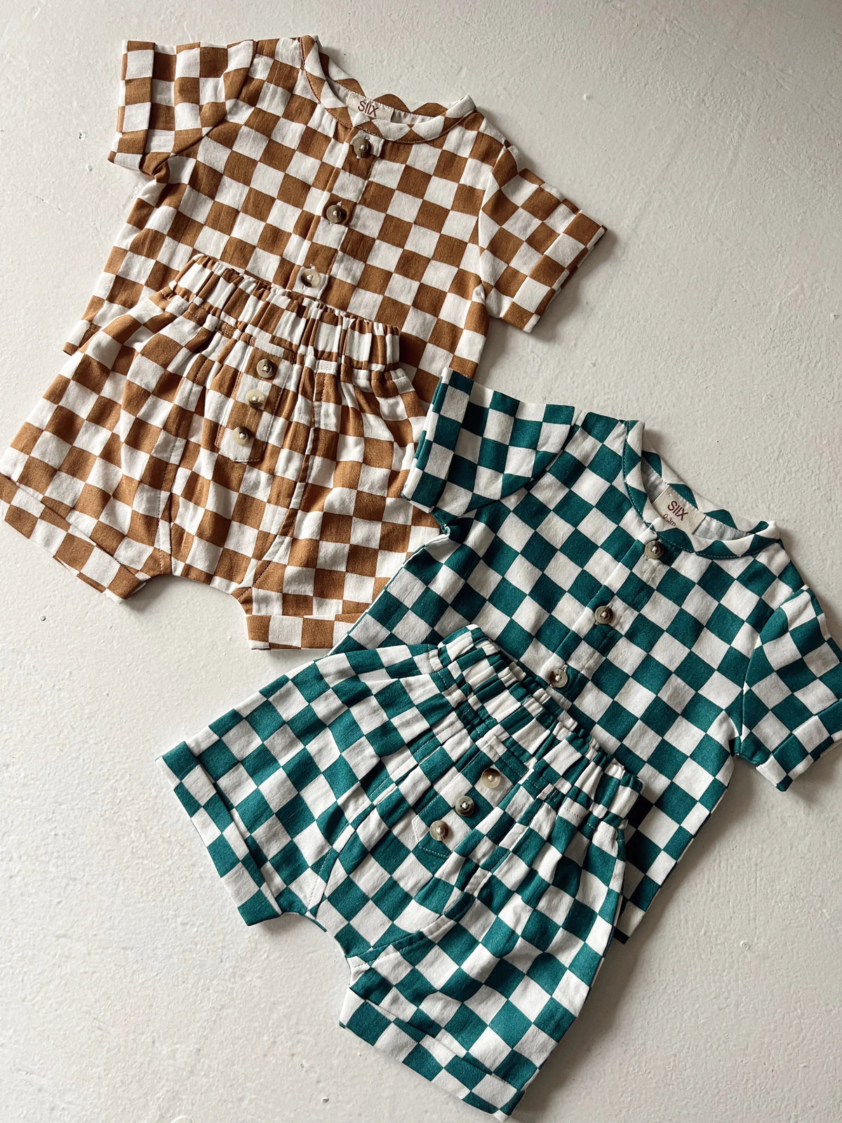 Two checkered rompers in brown and teal colors, featuring short sleeves and button details, displayed on a neutral background.