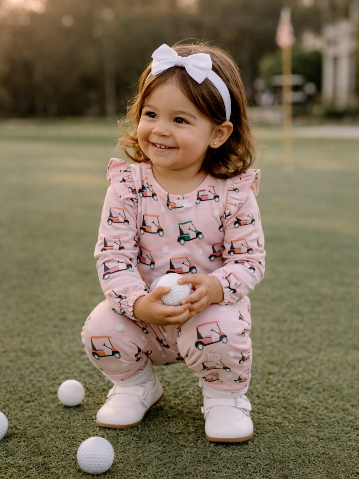 Smiling toddler in pink golf cart pajamas holds a golf ball on a green lawn, with sunshine in the background.