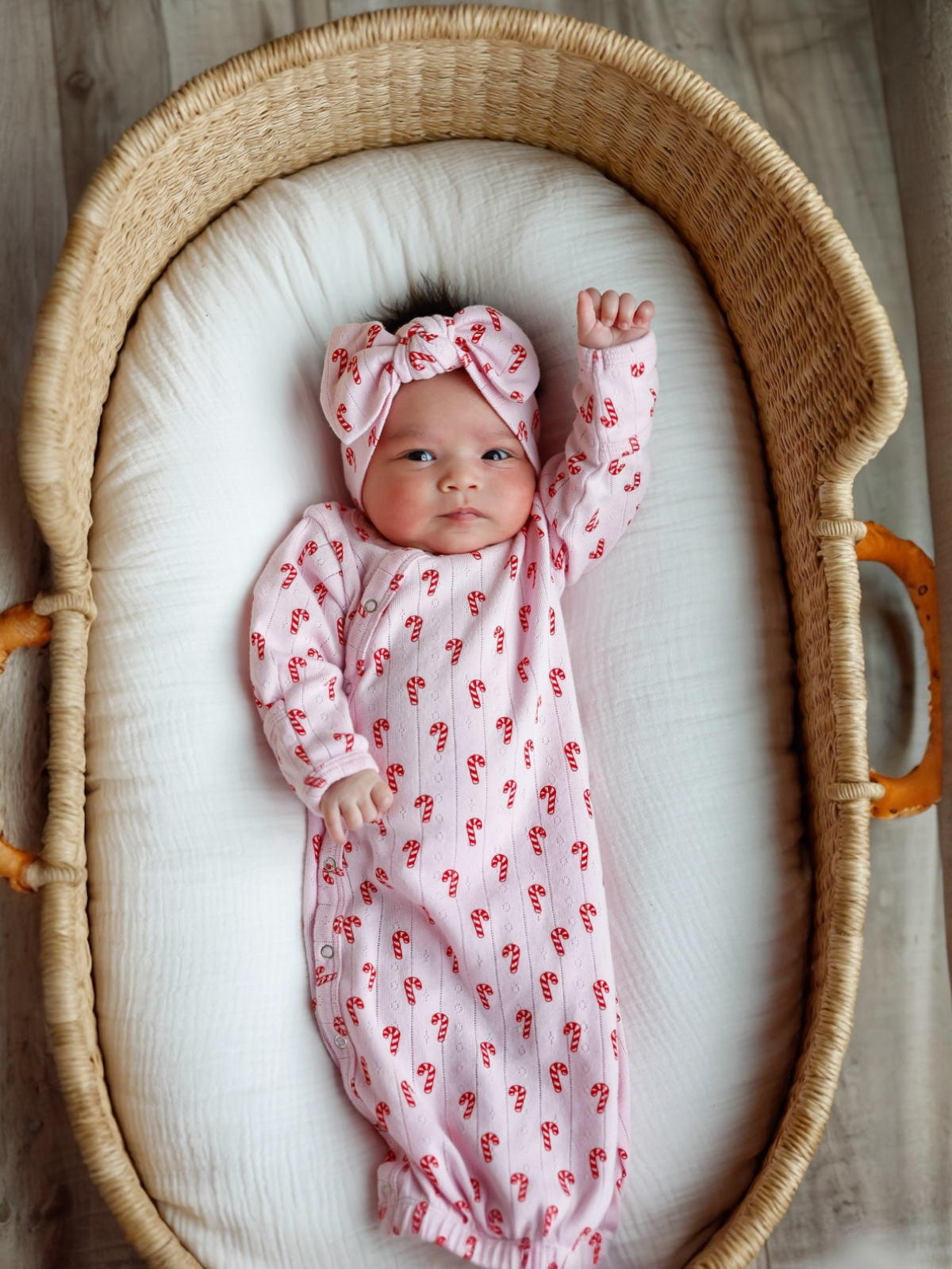 Infant in a pink candy cane patterned onesie and bow, lying in a woven basket on a soft white blanket.