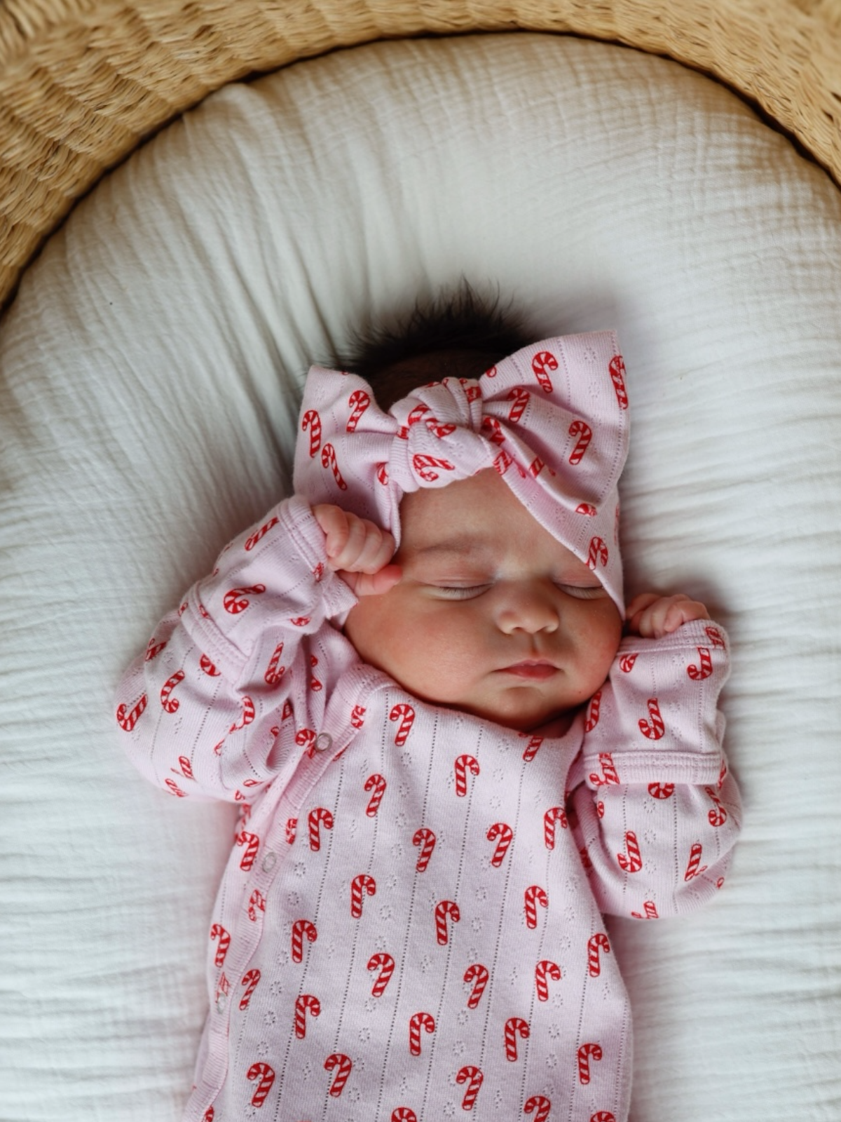 Newborn baby girl sleeping on a white cushion, wearing a pink outfit with candy cane pattern and a matching bow headband.