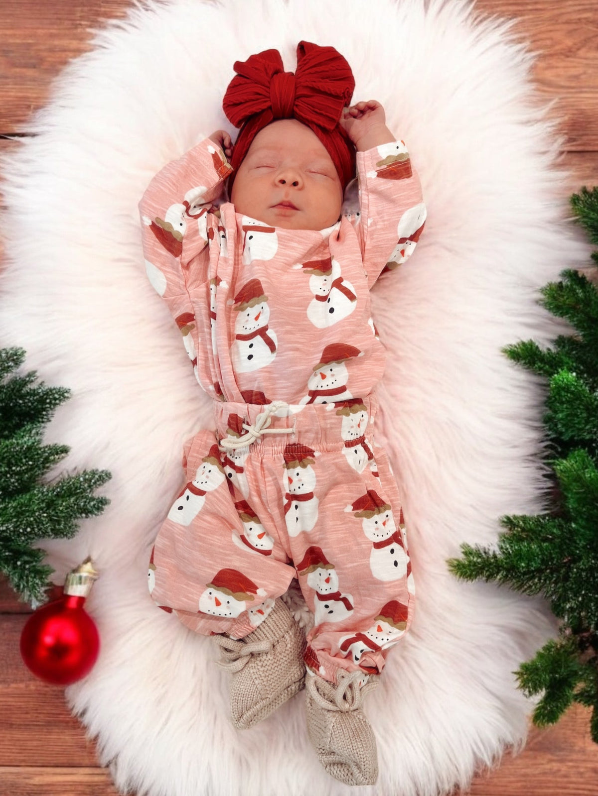 Baby asleep on a fluffy white rug, wearing a snowman-patterned outfit and a red bow headband, with festive decorations.