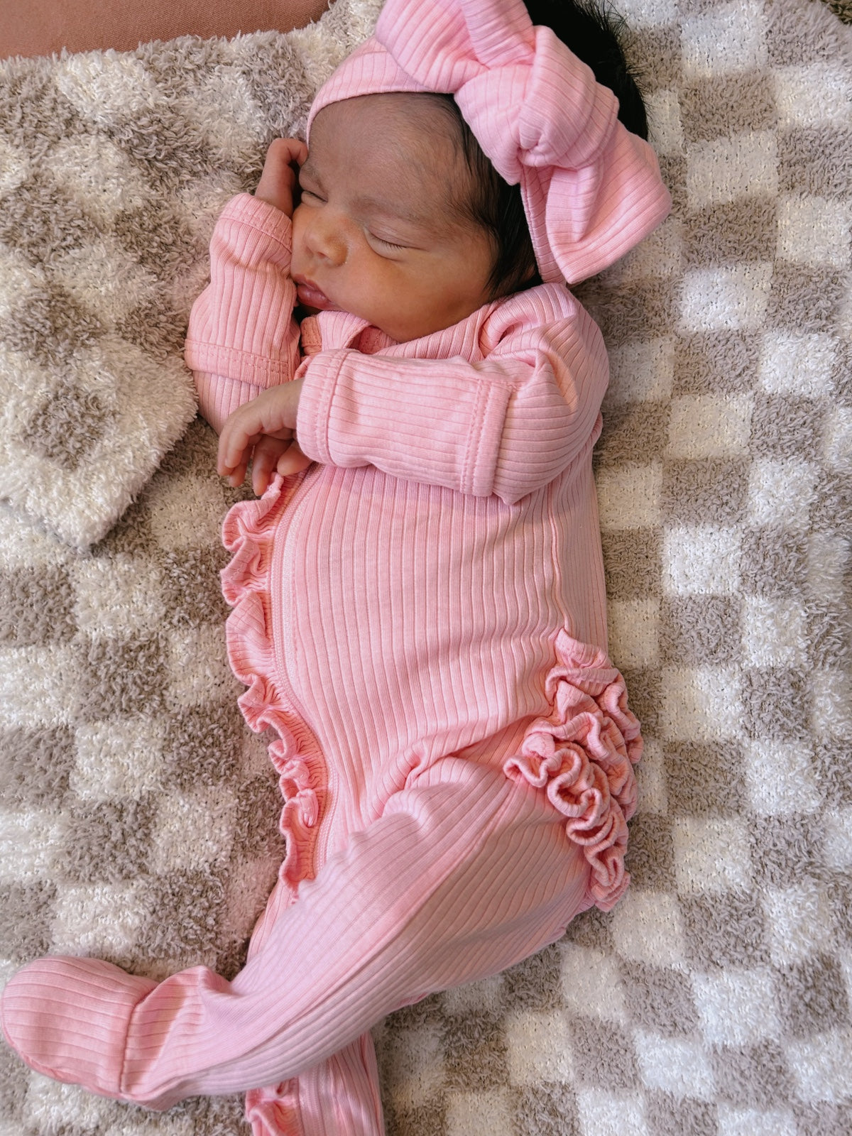 Newborn in a pink outfit with ruffles, sleeping on a soft checkered blanket with a large bow on her head.