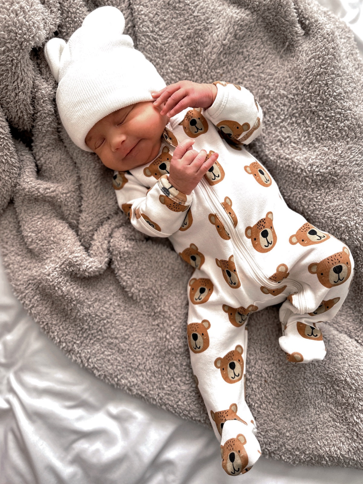 Infant wearing a bear-patterned outfit and white hat, smiling on a fluffy gray blanket.