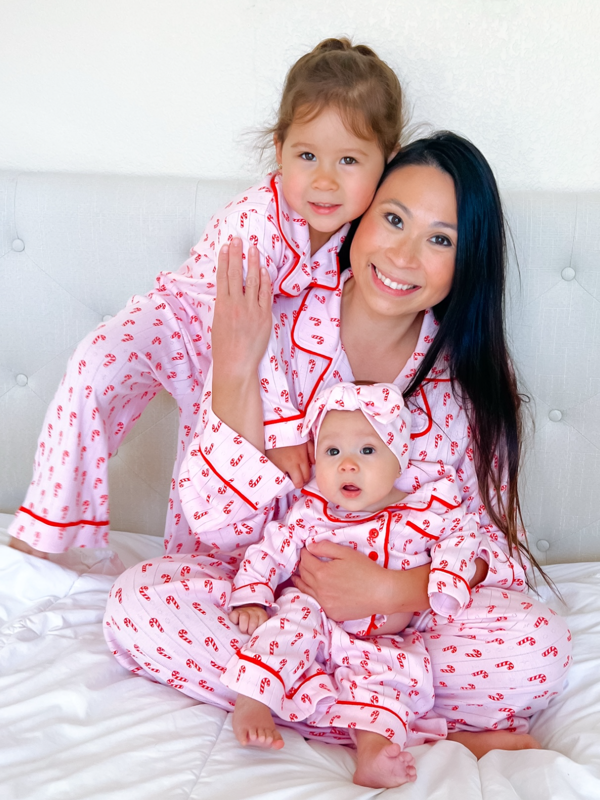 Mother and two daughters in matching pink pajamas with candy cane print, smiling together on a white bed.