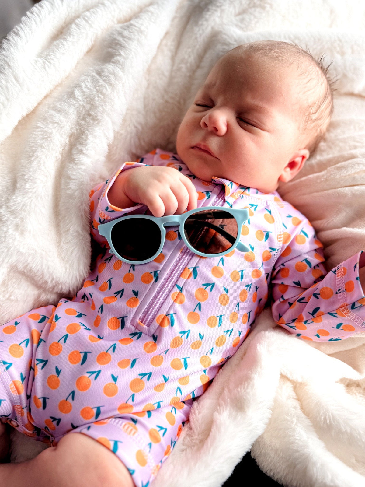 A sleeping baby in bright orange-patterned swimwear holds sunglasses while resting on a soft blanket.