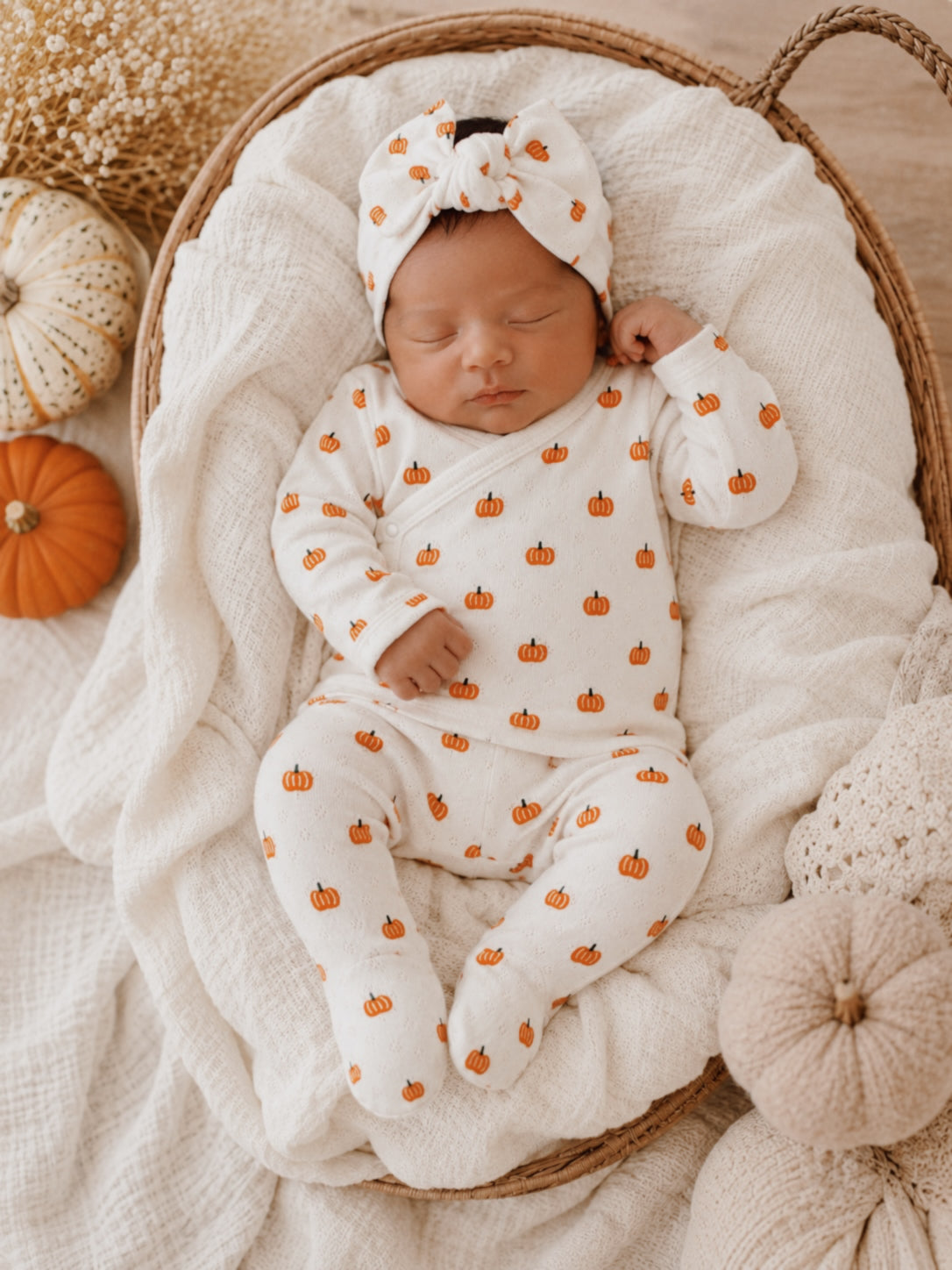 Newborn baby asleep in a cozy basket, wearing a pumpkin-patterned outfit and matching headband.