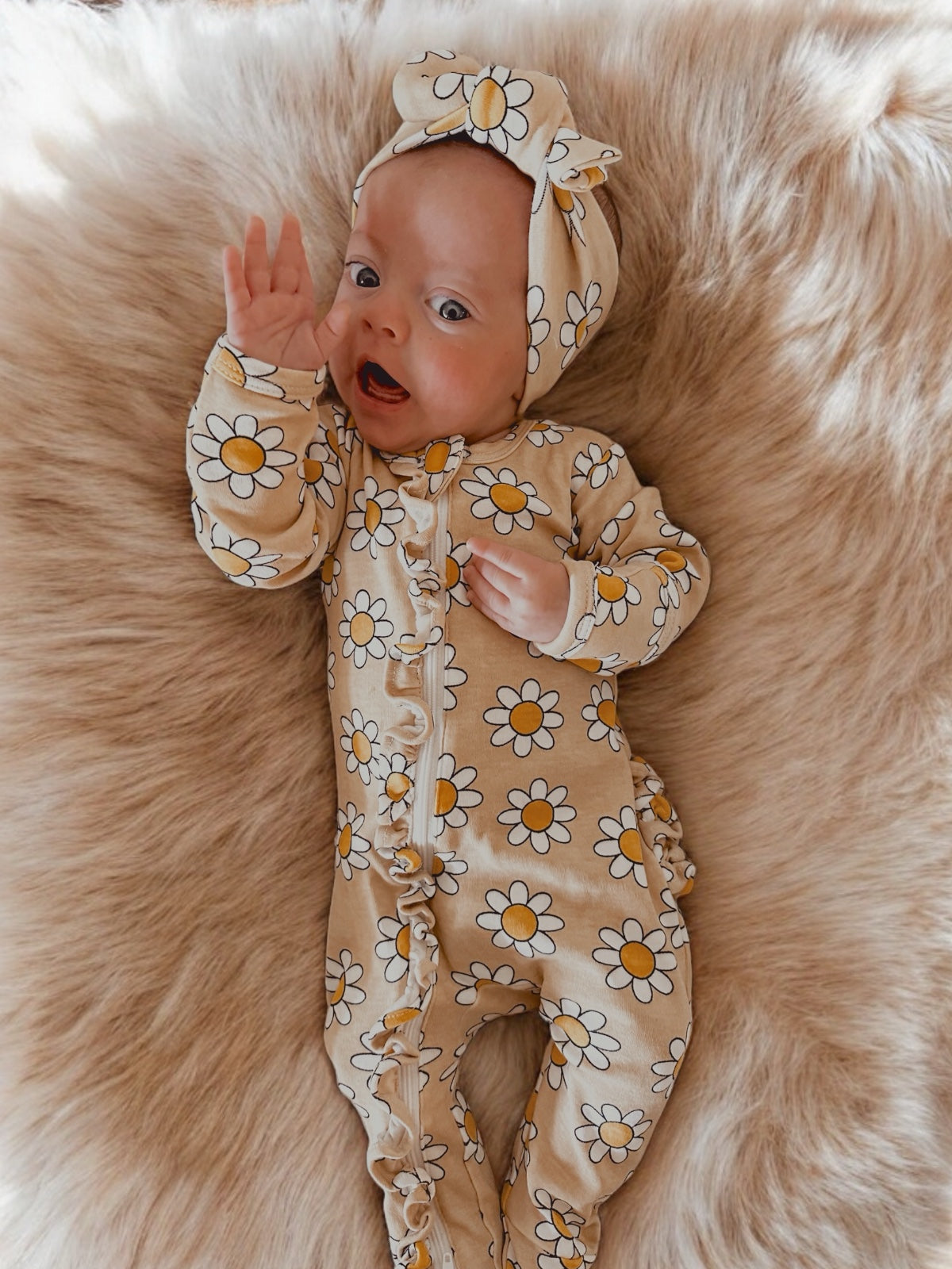 Infant wearing a floral onesie and headband, smiling and waving on a fluffy surface.