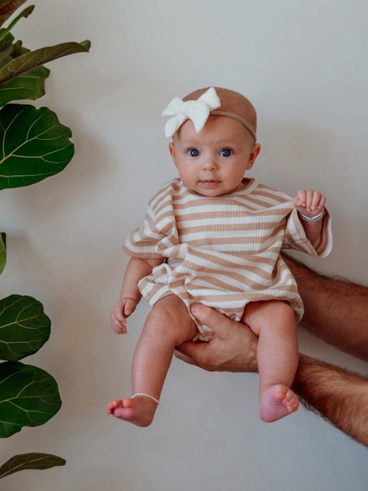 Baby in a striped outfit and bow, held by an adult hand with green leaves in the background.