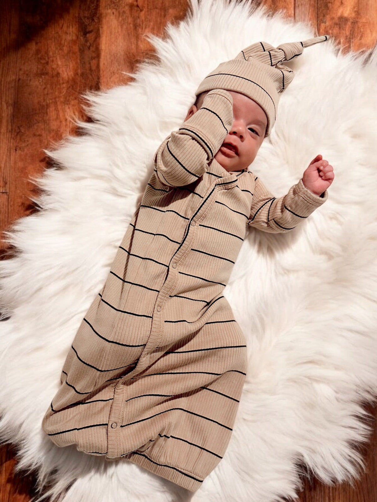 Infant wearing a striped tan outfit and hat, resting on a white fluffy rug.