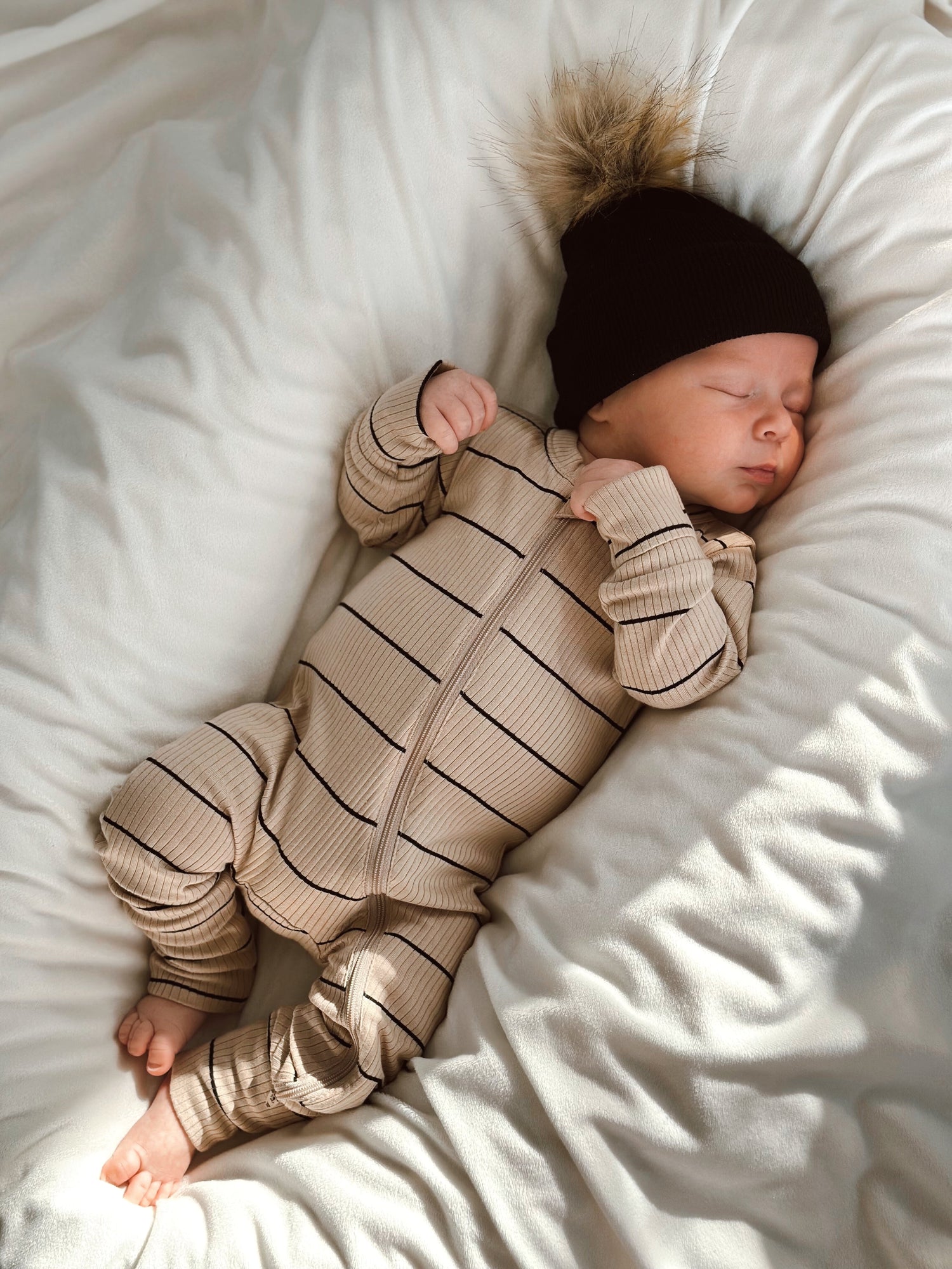 A peacefully sleeping baby in a striped onesie and a black hat with a pom-pom, resting on a soft white blanket.