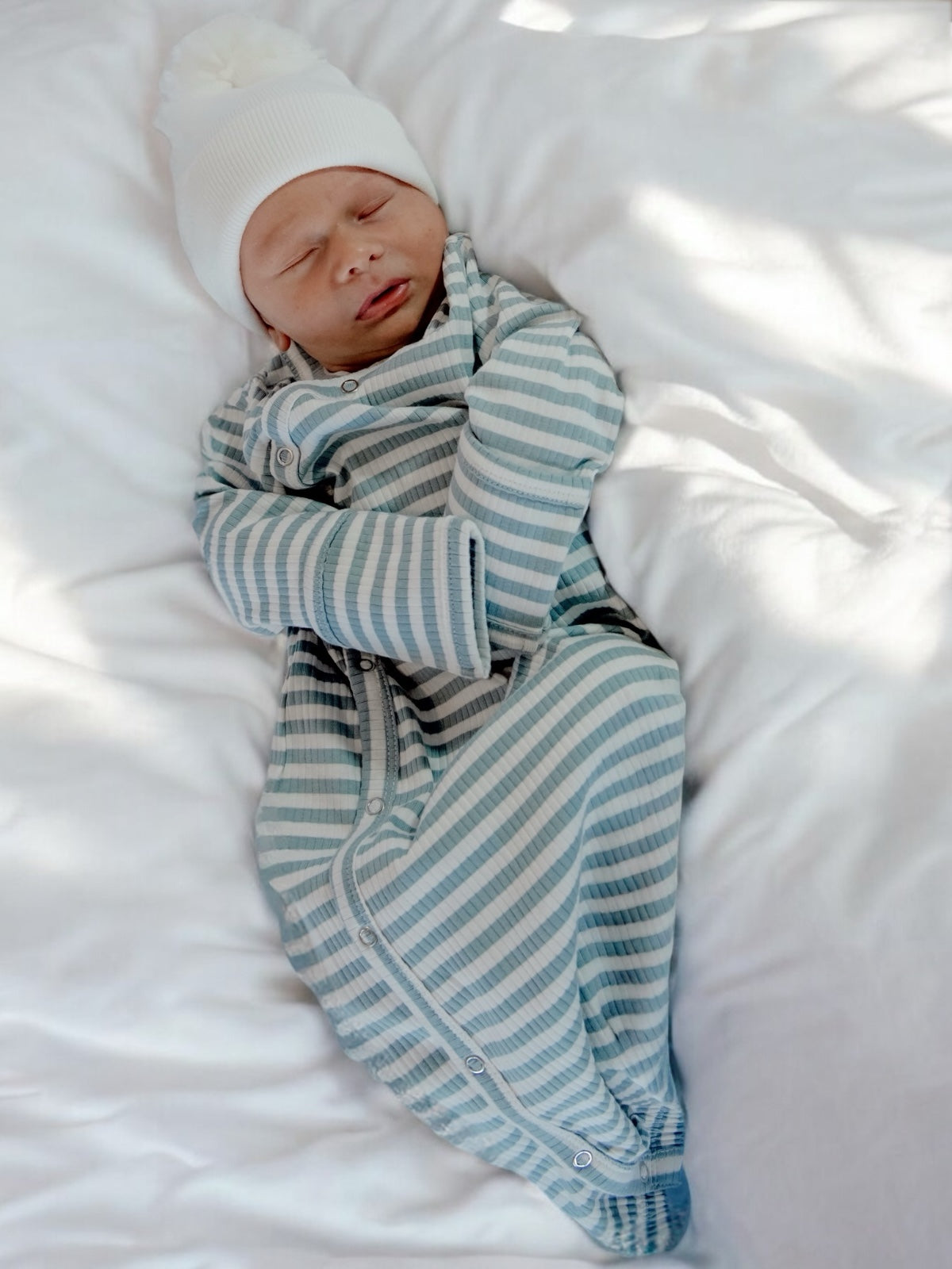 Sleeping baby in striped pajamas and white hat, relaxed on a soft bed.
