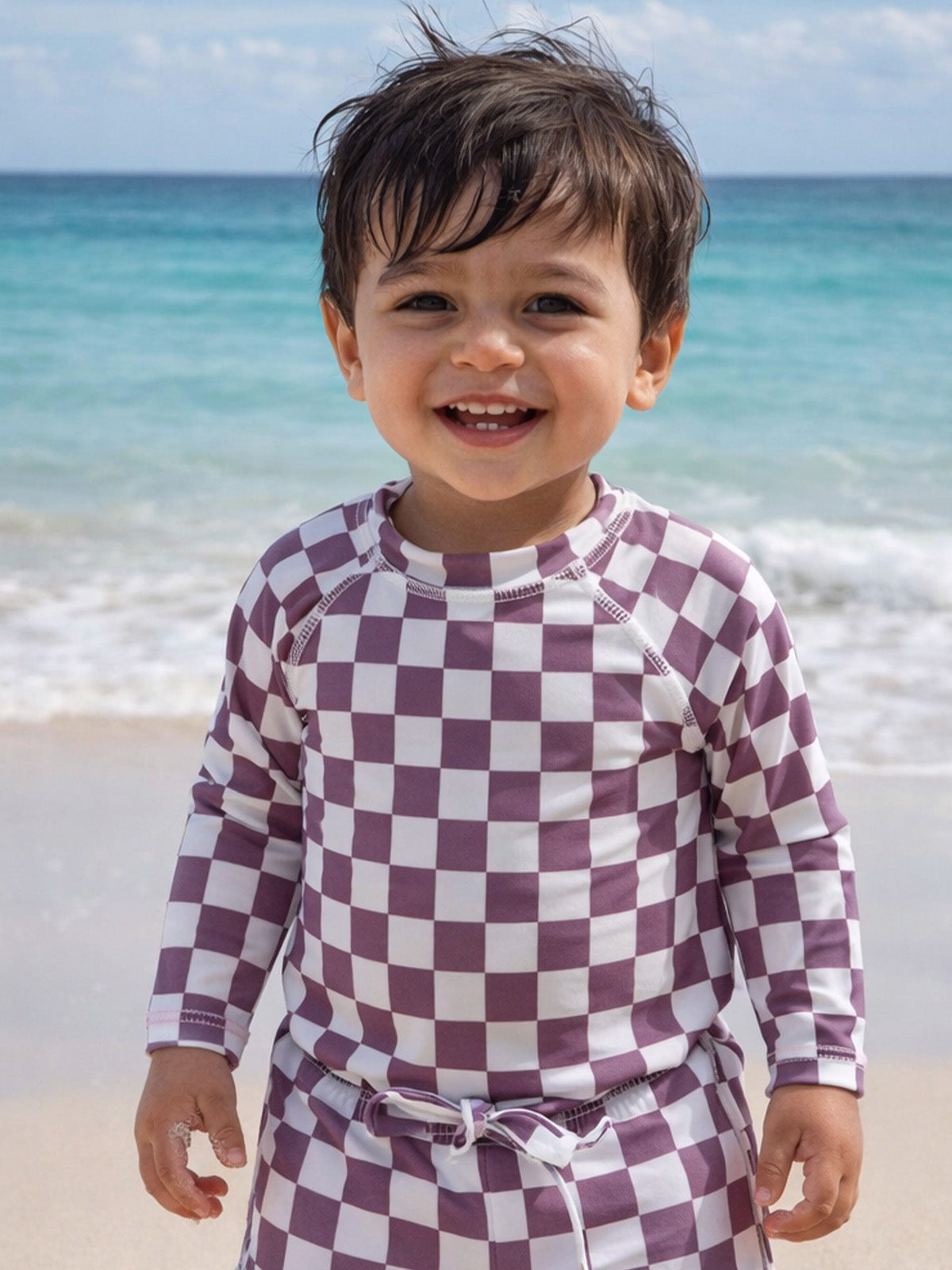Smiling toddler in a checkered swimsuit on the beach, with ocean waves in the background.