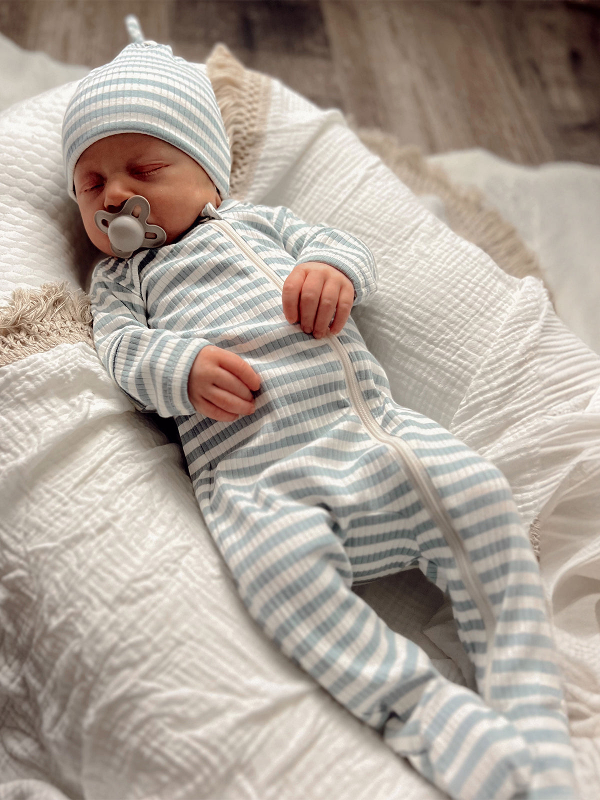 Infant in blue-striped outfit and hat, sleeping peacefully on a textured white blanket.