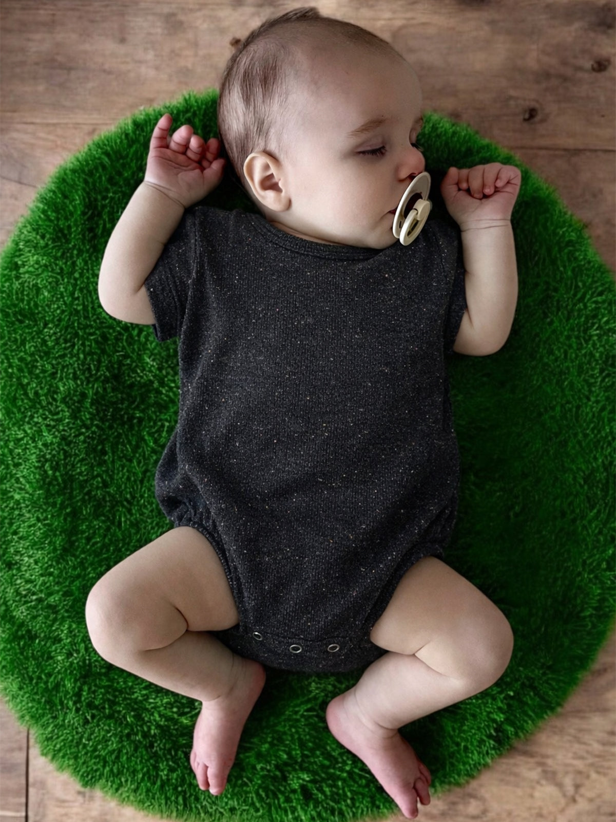 Infant sleeping peacefully on a green rug, wearing a dark onesie and a pacifier, with arms relaxed and legs spread.