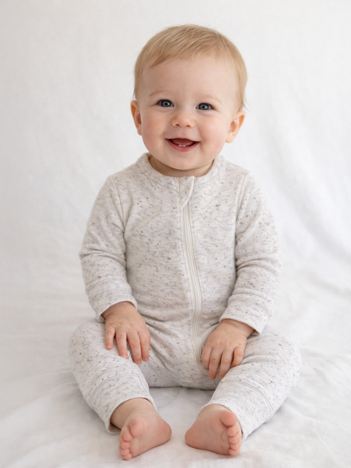 Smiling baby in a light gray onesie, sitting on white fabric, showcasing joy and warmth.