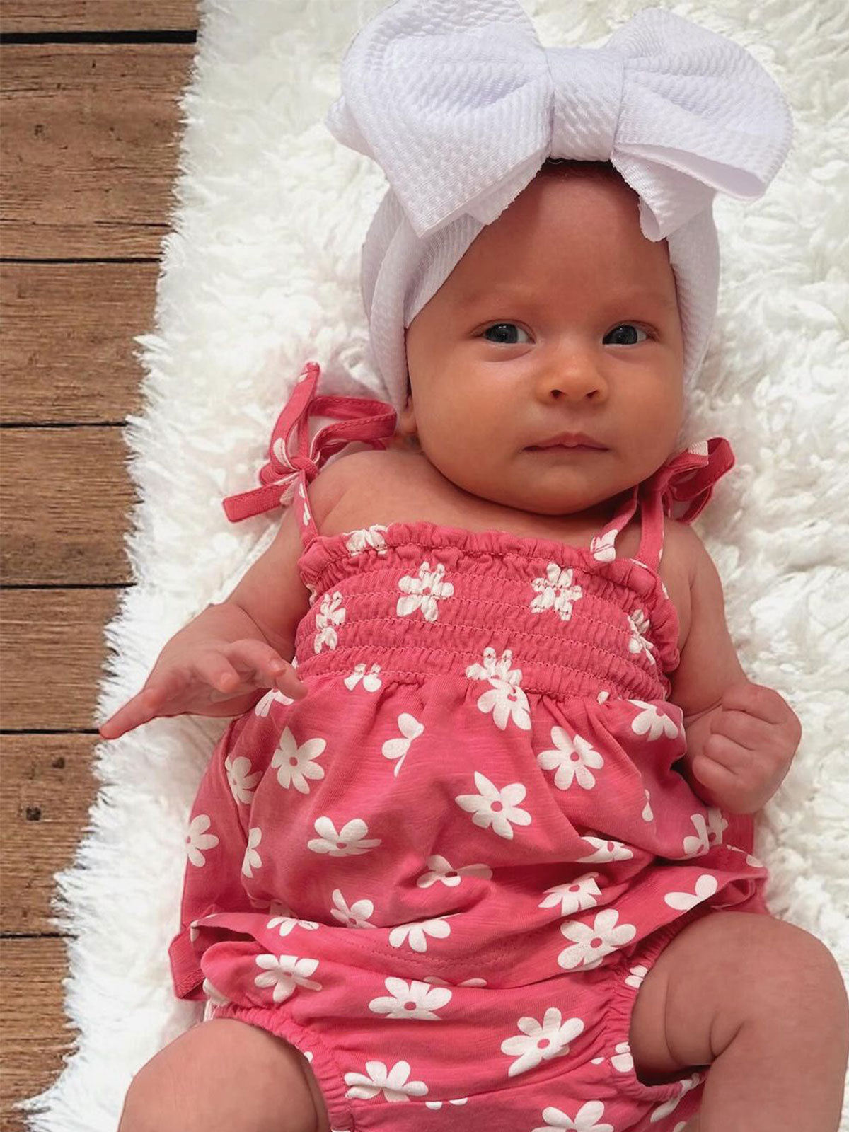 Infant wearing a pink floral outfit and a large white bow, lying on a soft white blanket.