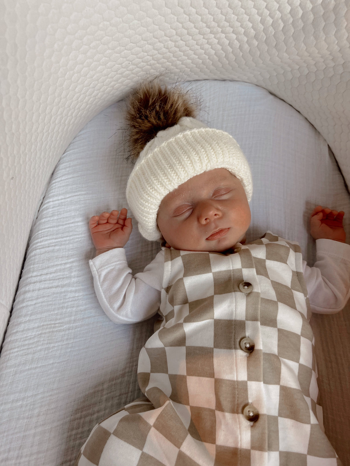 Sleeping baby in a cozy white hat on a textured blanket, dressed in a checkered outfit.