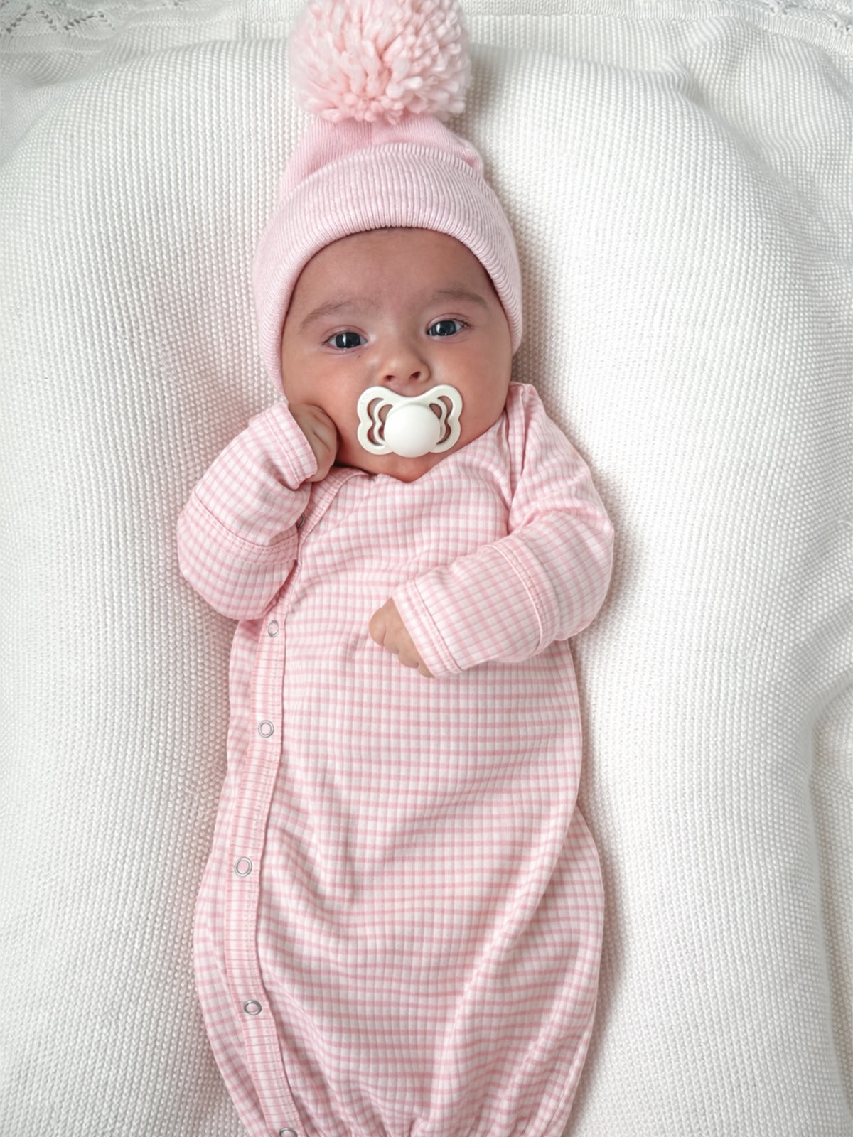 Smiling baby in a pink checkered outfit and pom-pom hat, sucking a pacifier on a textured blanket.