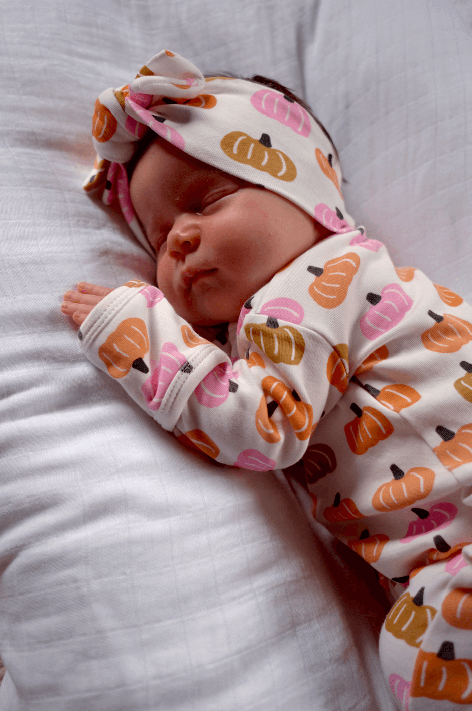 Baby sleeping peacefully on a white pillow, wearing a pumpkin-patterned onesie and matching headband.