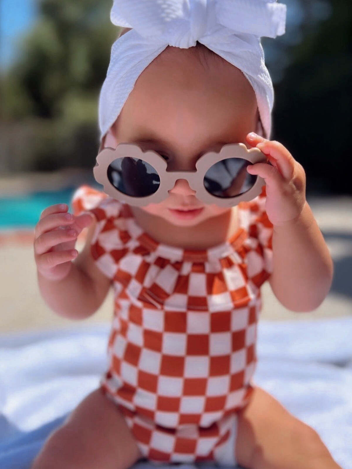 Baby wearing a checkered swimsuit and sunglasses, sitting on a blanket by the pool.