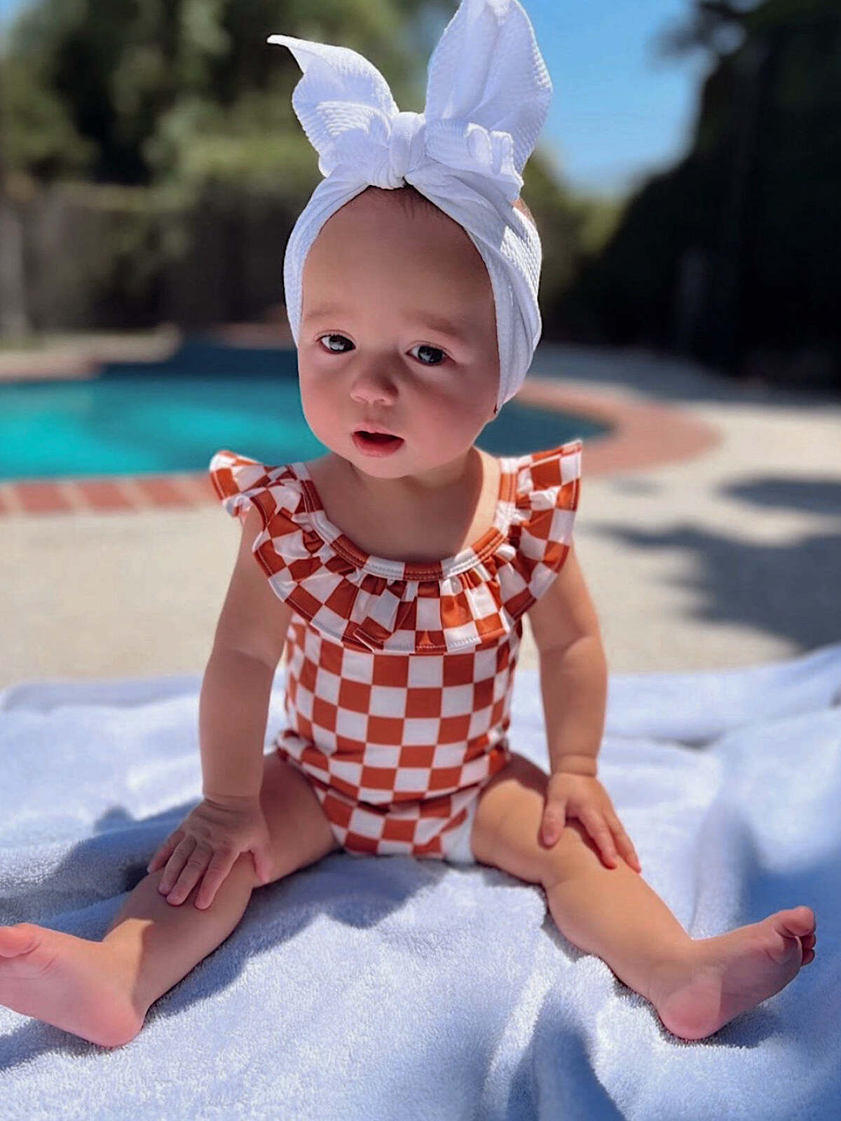Baby in a checkered swimsuit and white bow sits on a towel by a poolside on a sunny day.