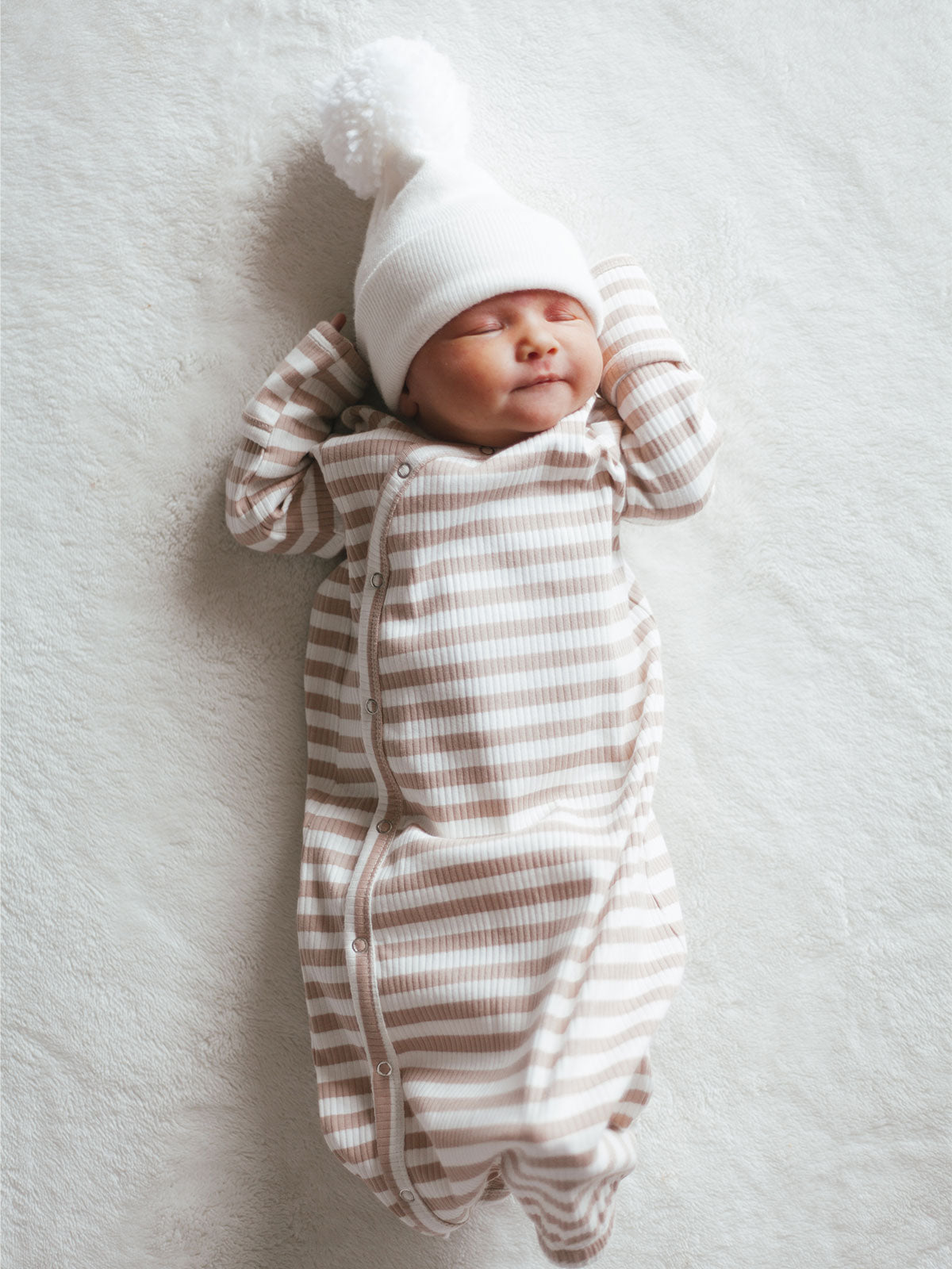 Smiling newborn in a striped onesie and pom-pom hat, laying on a soft blanket.