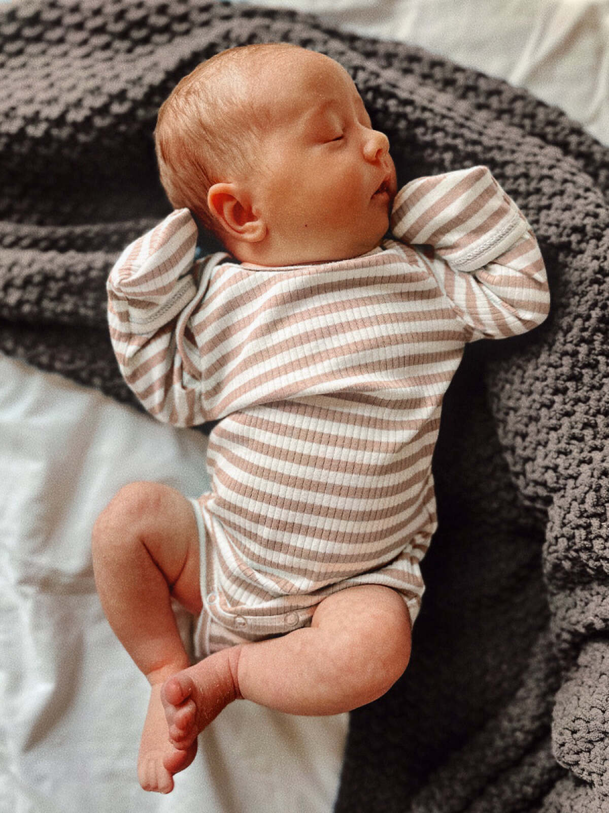 Sleepy baby in striped onesie, nestled on a textured blanket, with arms raised and eyes closed.