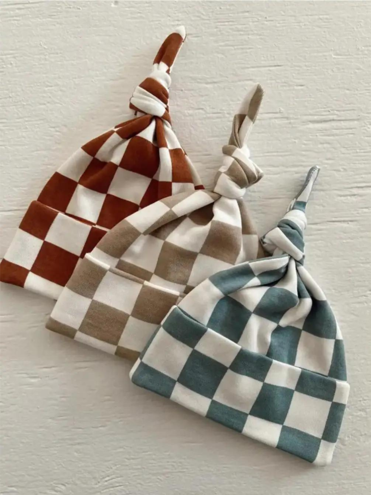 Three stylish checkered baby hats in rust, beige, and teal on a textured white background.